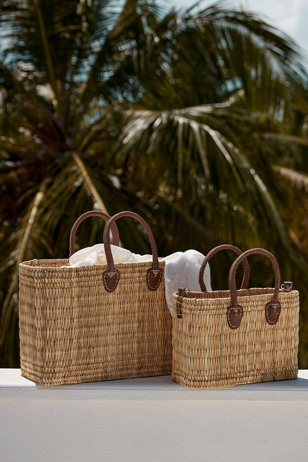 Two woven straw tote bags with leather handles and white cloths, set on a white ledge with palm fronds in the background.