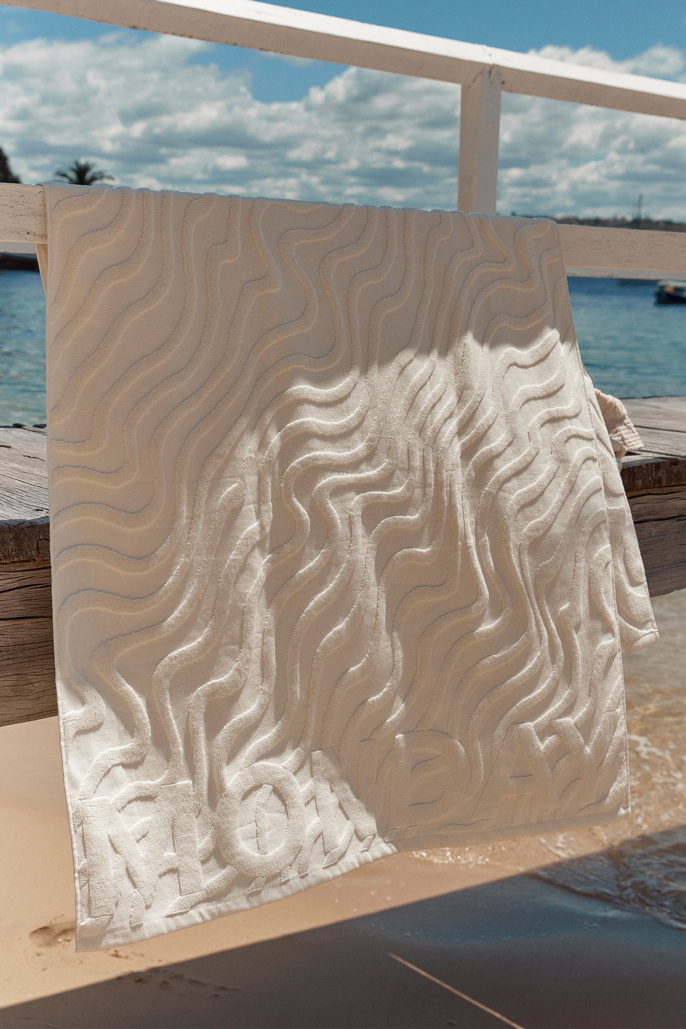 Beige towel with raised wavy texture draped over a pier railing, embossed MONACO, sea and clouds behind.