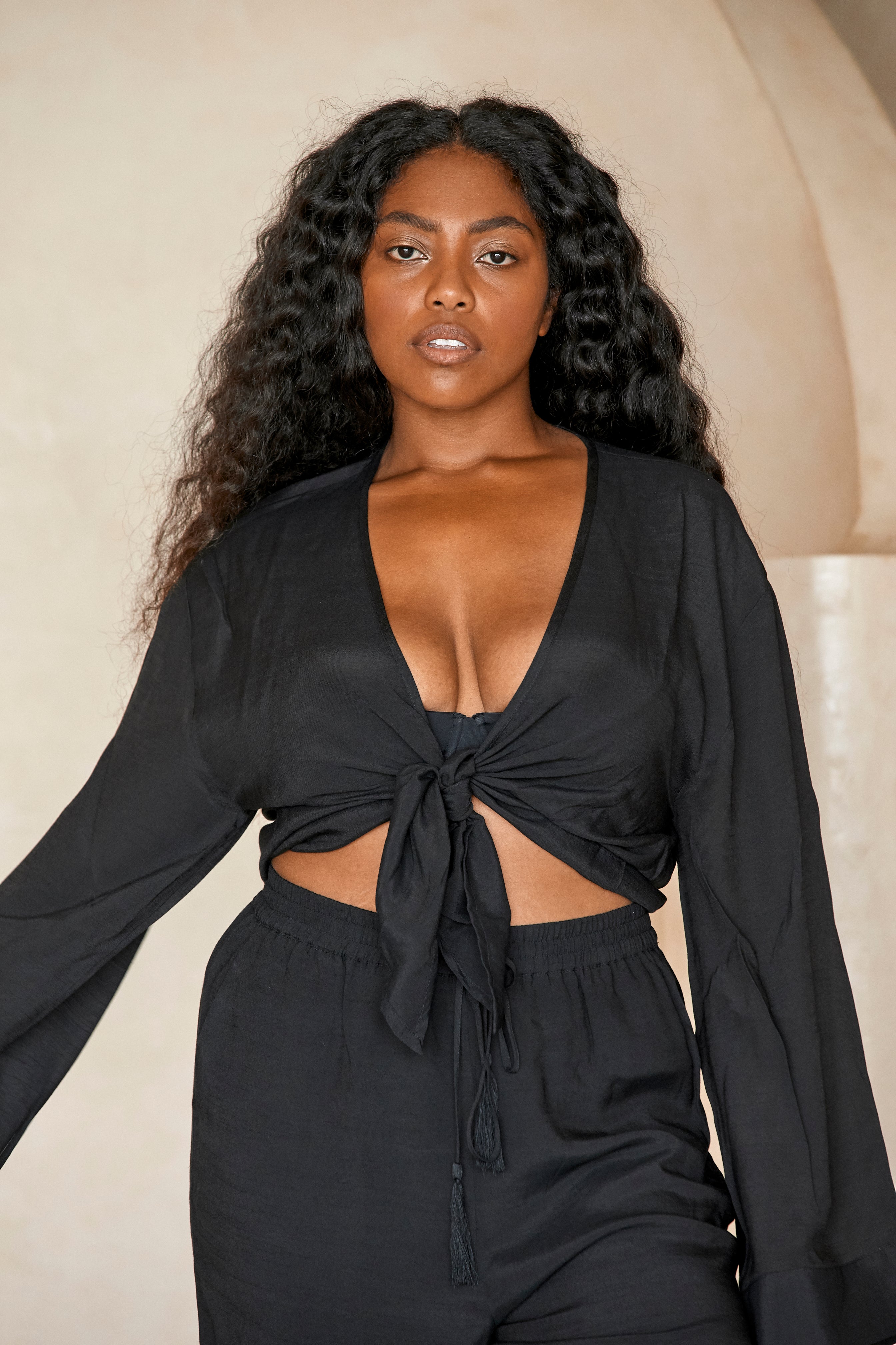 Woman with long curly hair wearing black tie-front cropped top and high-waist drawstring pants, neutral studio background.