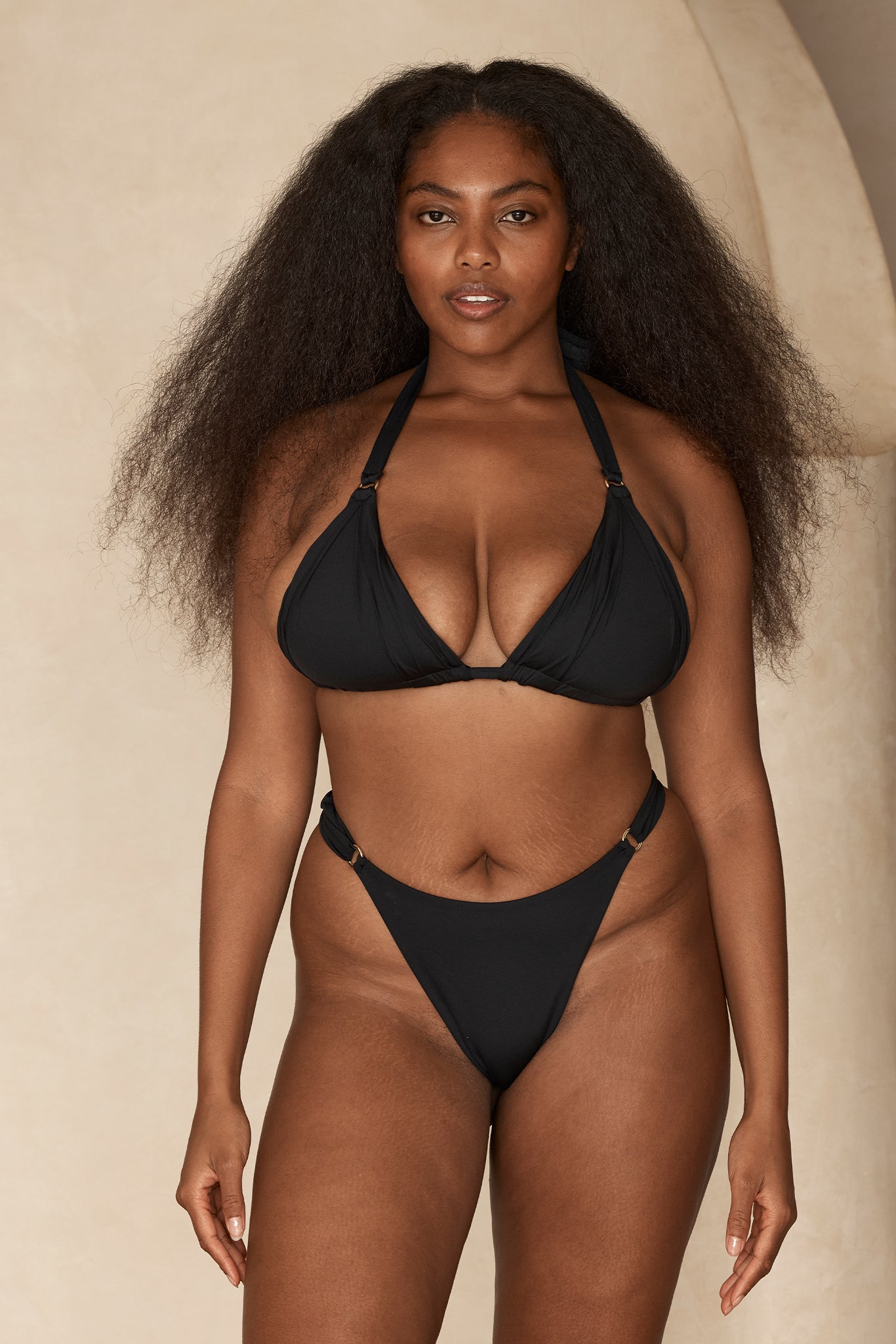 Front-facing Black woman with long natural hair wearing a black bikini against a neutral studio background.