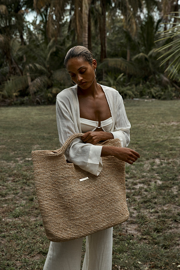 Woman in white outfit holding a large woven straw tote bag in a grassy tropical setting