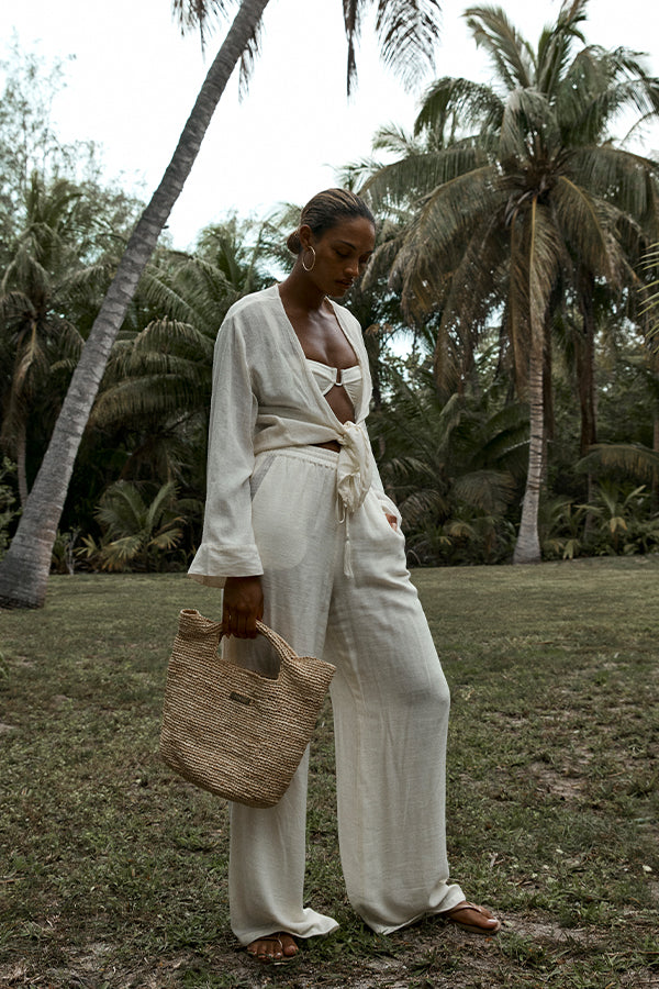 Woman in cream linen wrap top and wide pants holding a woven straw tote under palm trees