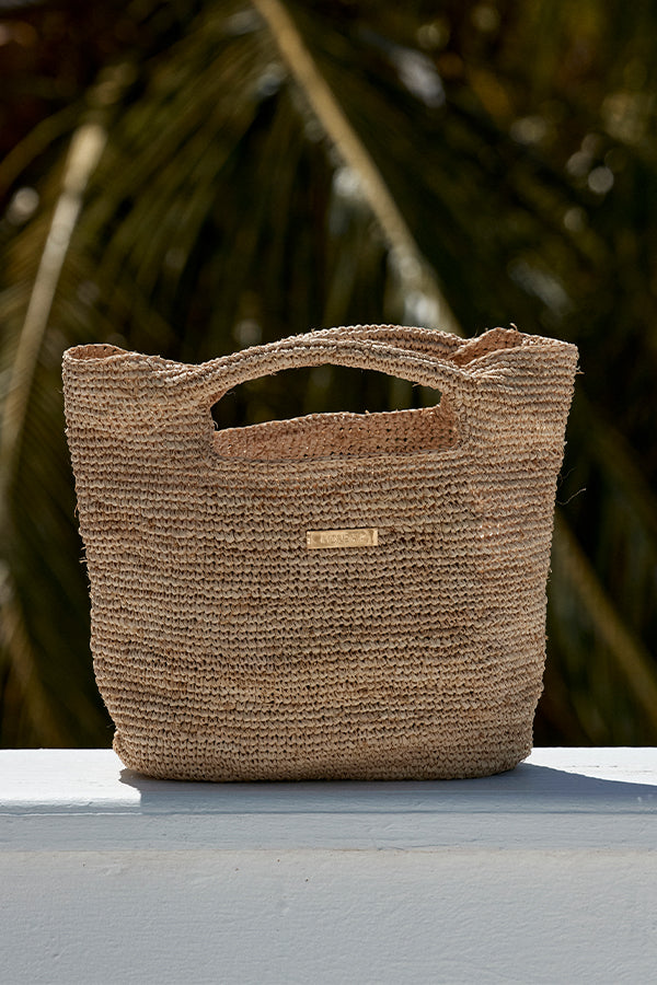 Woven straw tote bag with cutout handles on a white ledge and blurred palm-frond background.