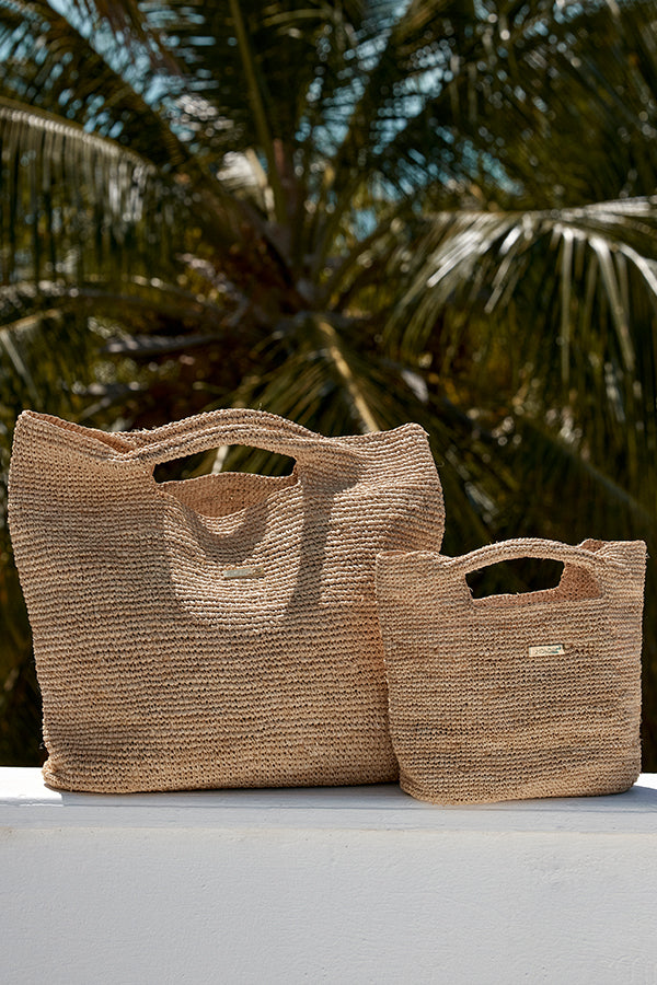 Two woven straw tote bags (large and small) with cutout handles on a white ledge, palm fronds in the background.