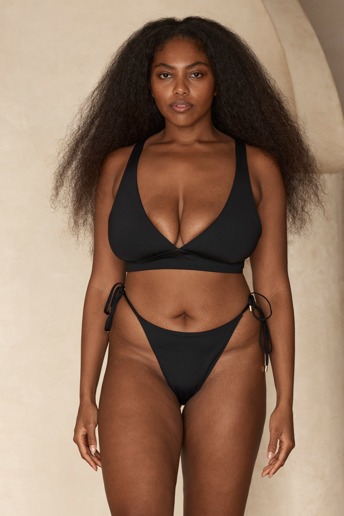 Front-facing model wearing a black V-neck bikini top and black tie-side bottoms, long voluminous hair, neutral backdrop.