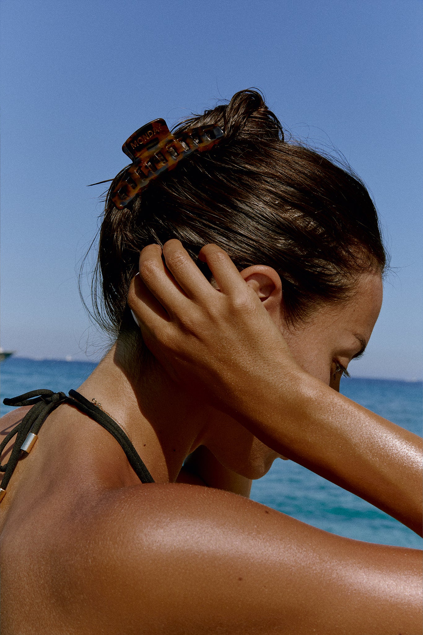 Close-up profile of a woman at the sea, adjusting hair secured with a tortoiseshell claw clip labeled MONDA.