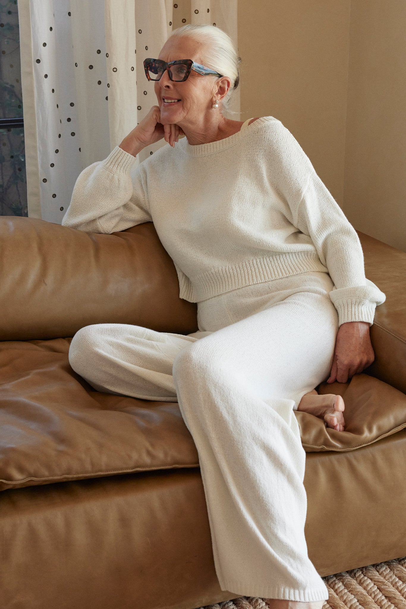 Older woman with white hair and tortoiseshell glasses sitting on tan leather sofa in cream knit sweater and pants, smiling.