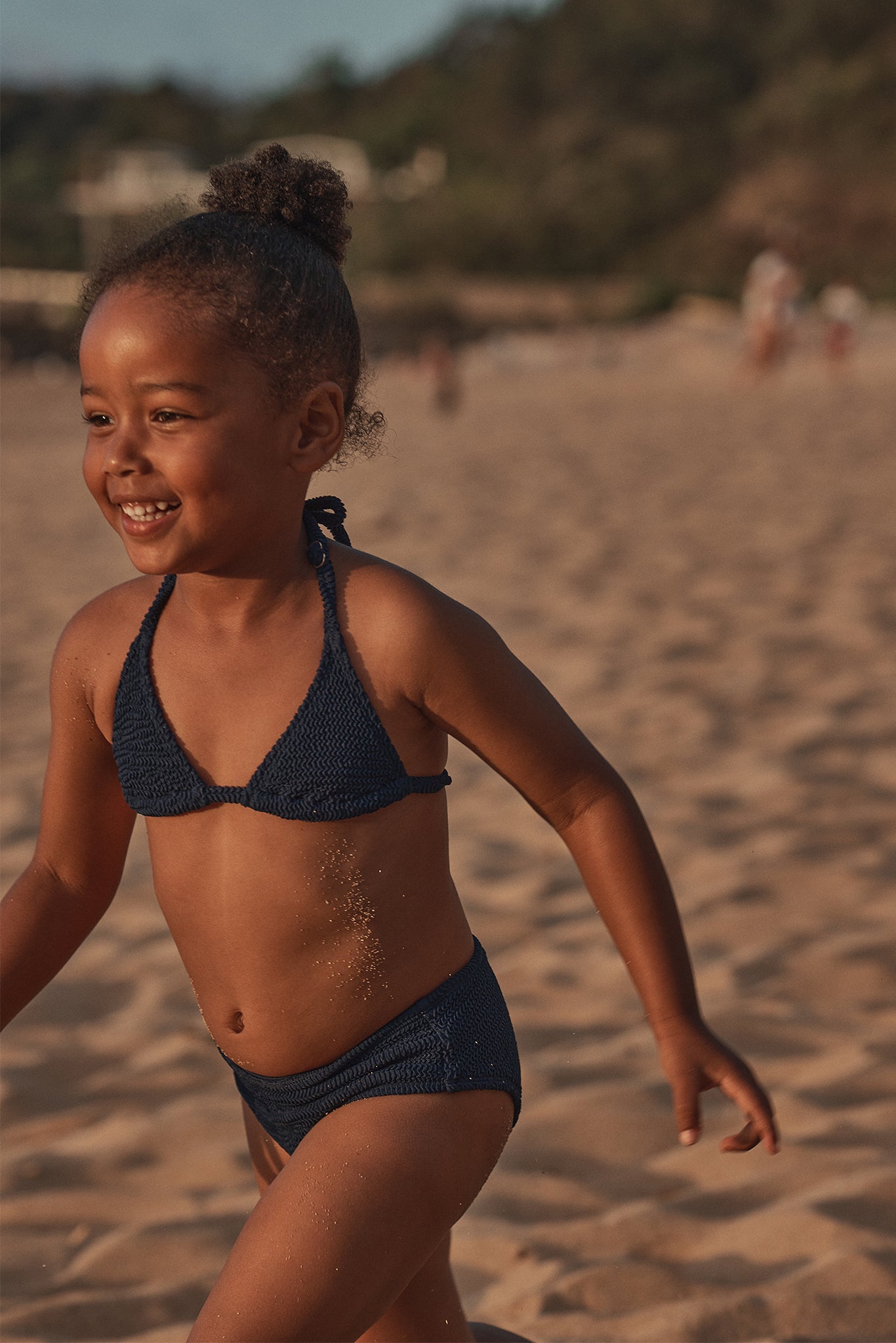 Young girl running on a sandy beach at sunset, smiling and wearing a navy knit bikini