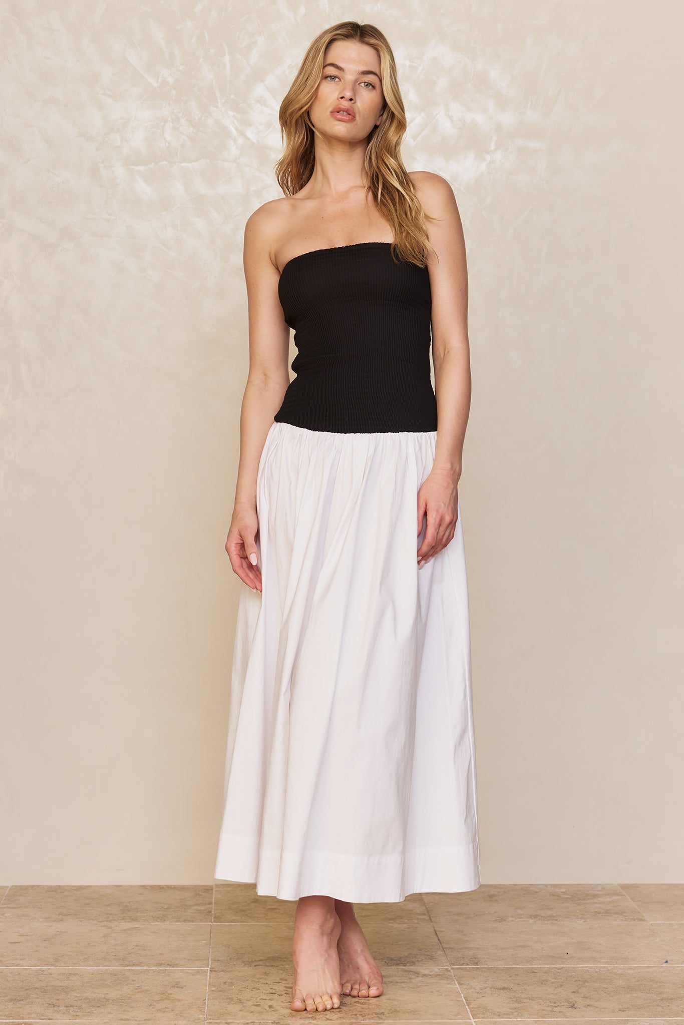 Woman standing barefoot in a black strapless top and long white skirt on neutral studio background