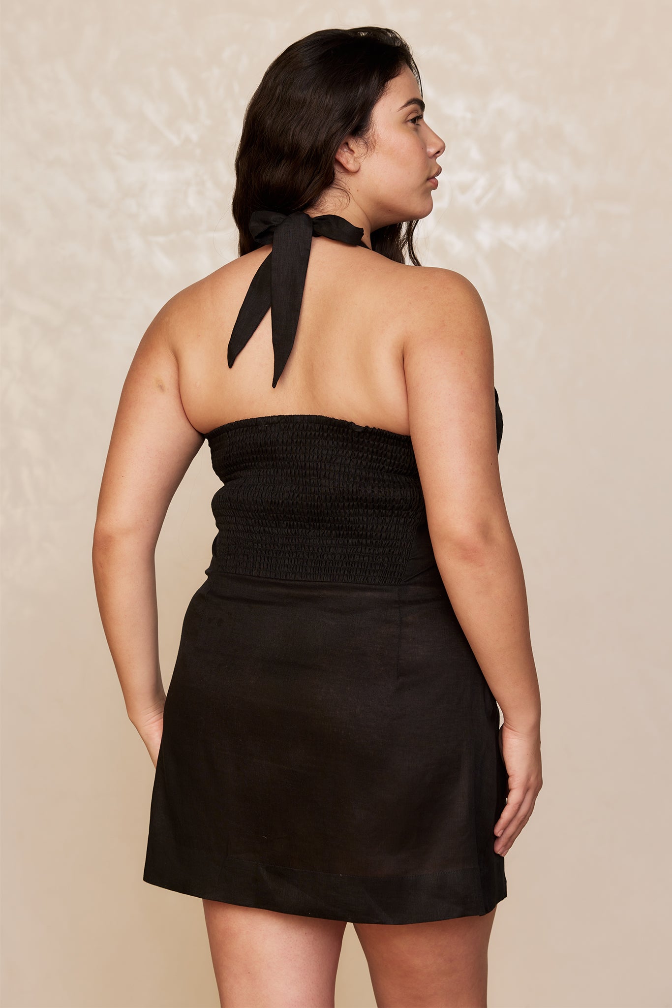 Back view of a woman wearing a black halter mini dress with smocked bodice and tied neck