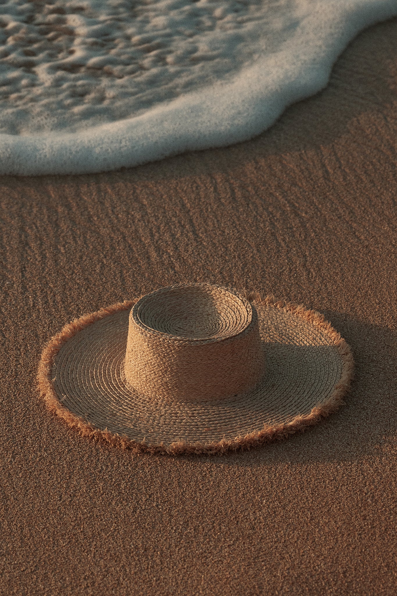 Straw sun hat resting on wet sand at the water's edge with a foamy wave