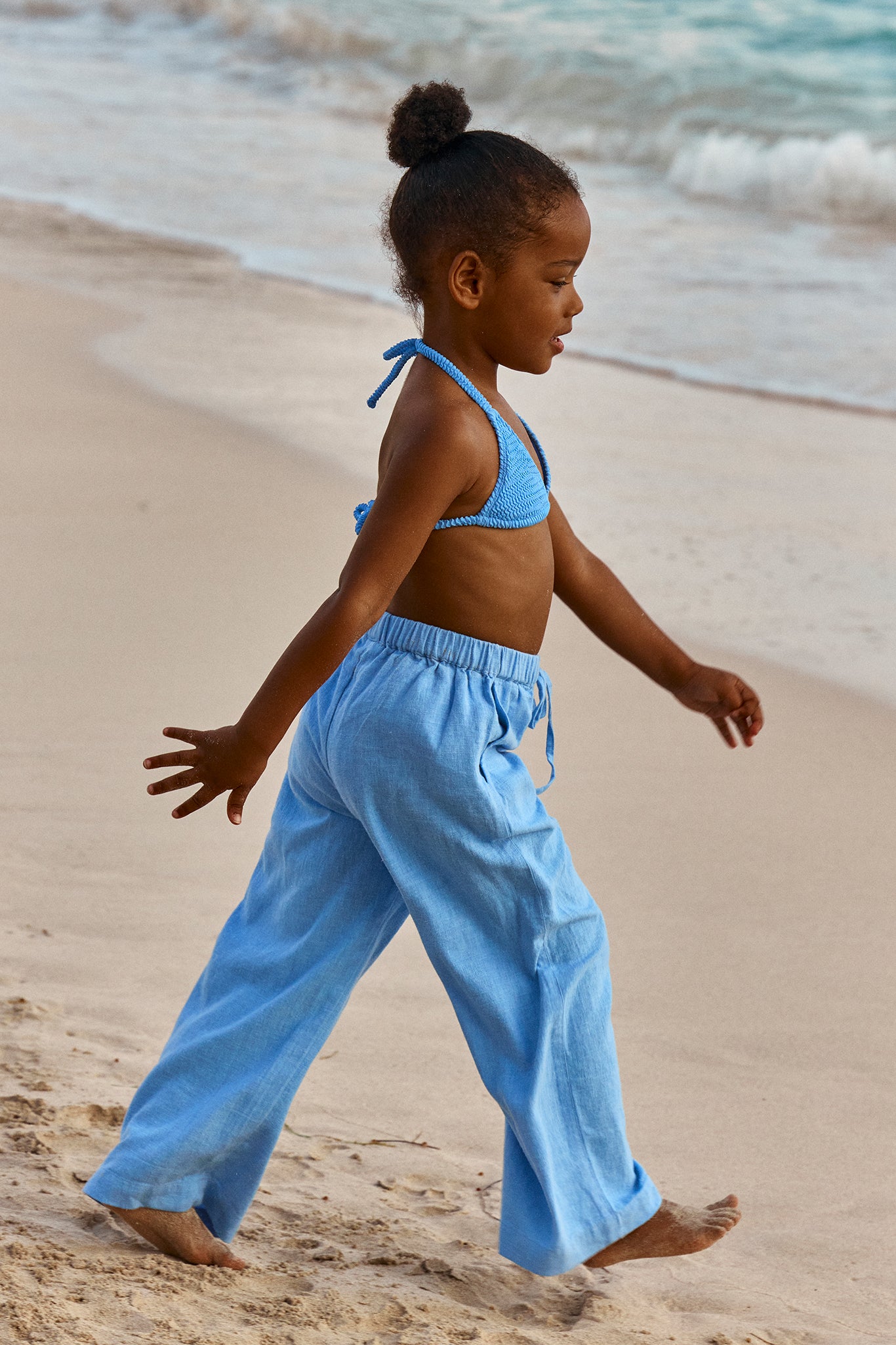 Young girl in a blue bikini top and loose light-blue pants walking barefoot along a sandy beach, side profile.