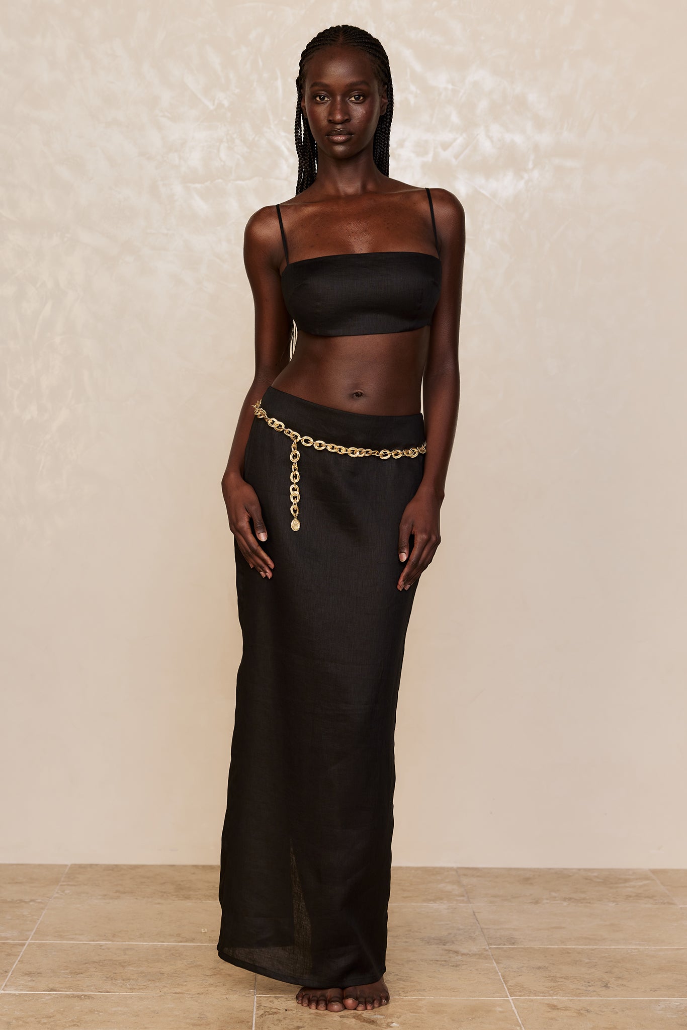 Tall dark-skinned model with braided hair wearing a black bandeau top and long black skirt with a gold chain belt, barefoot.