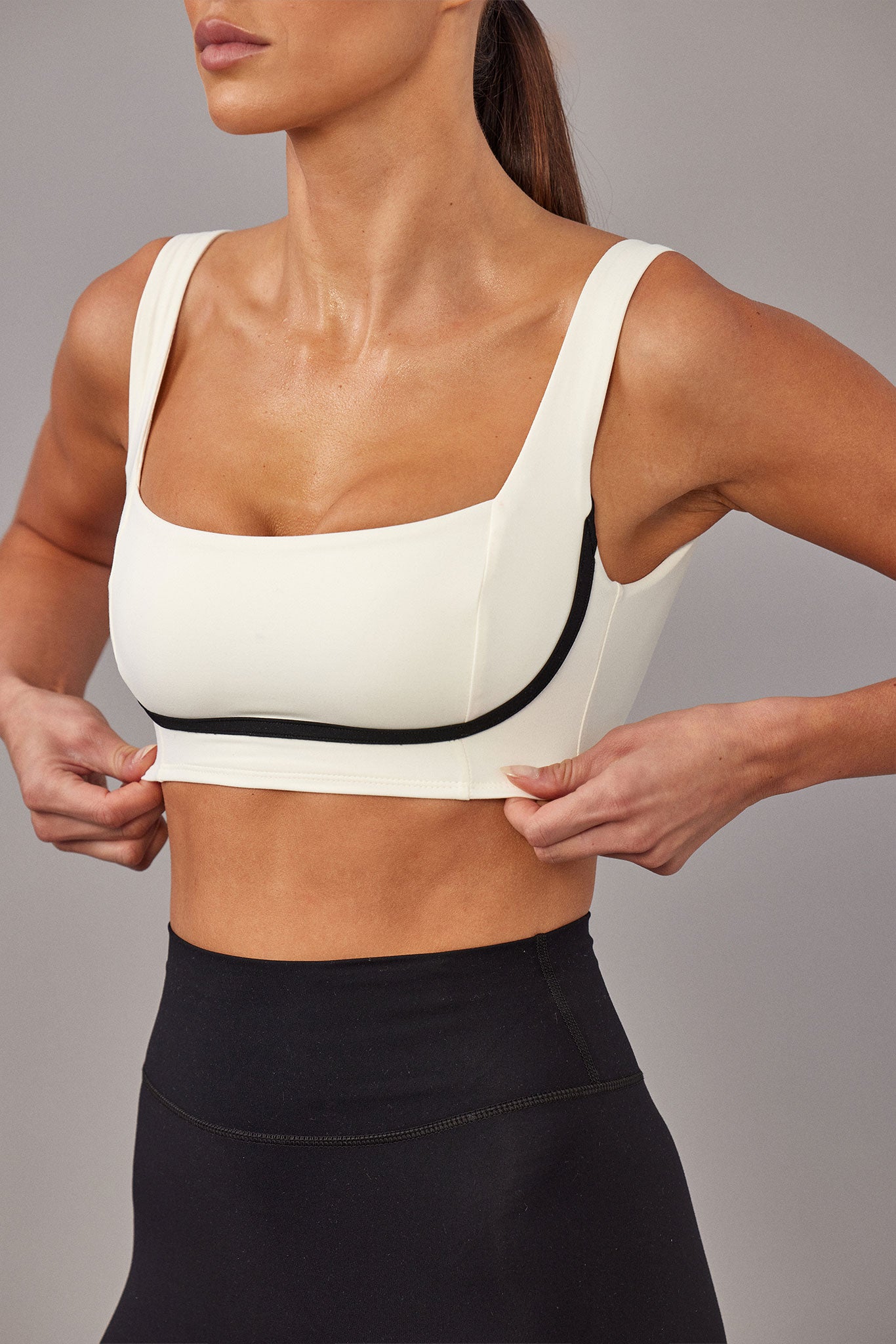 Athletic model adjusting a cream crop sports bra with black piping, shown from jaw to hips with high-waist black leggings.