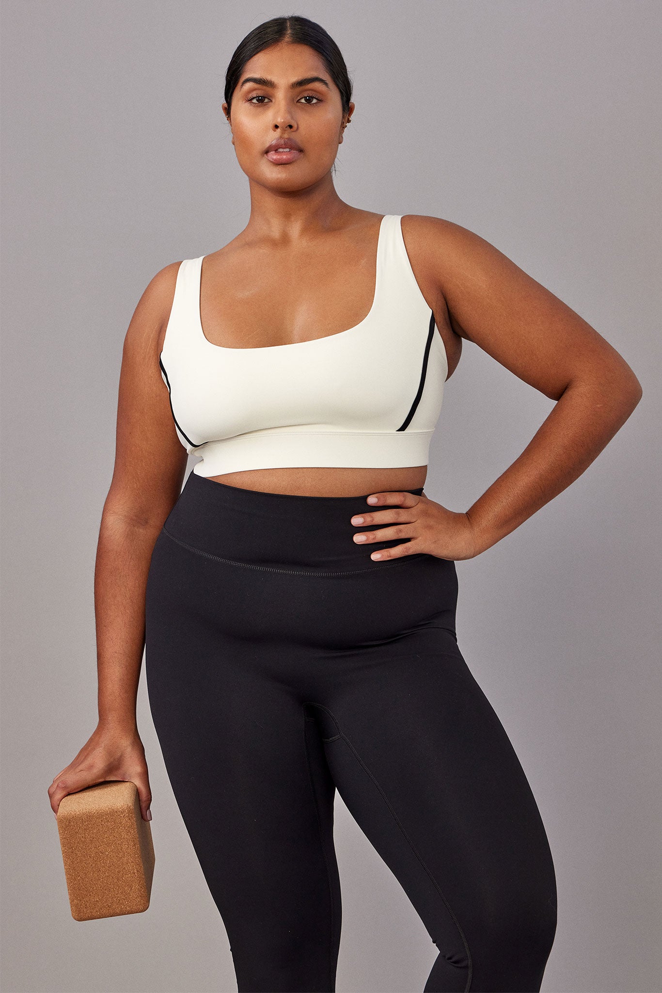 Plus-size model wearing cream sports bra and high-waist black leggings, holding a cork yoga block