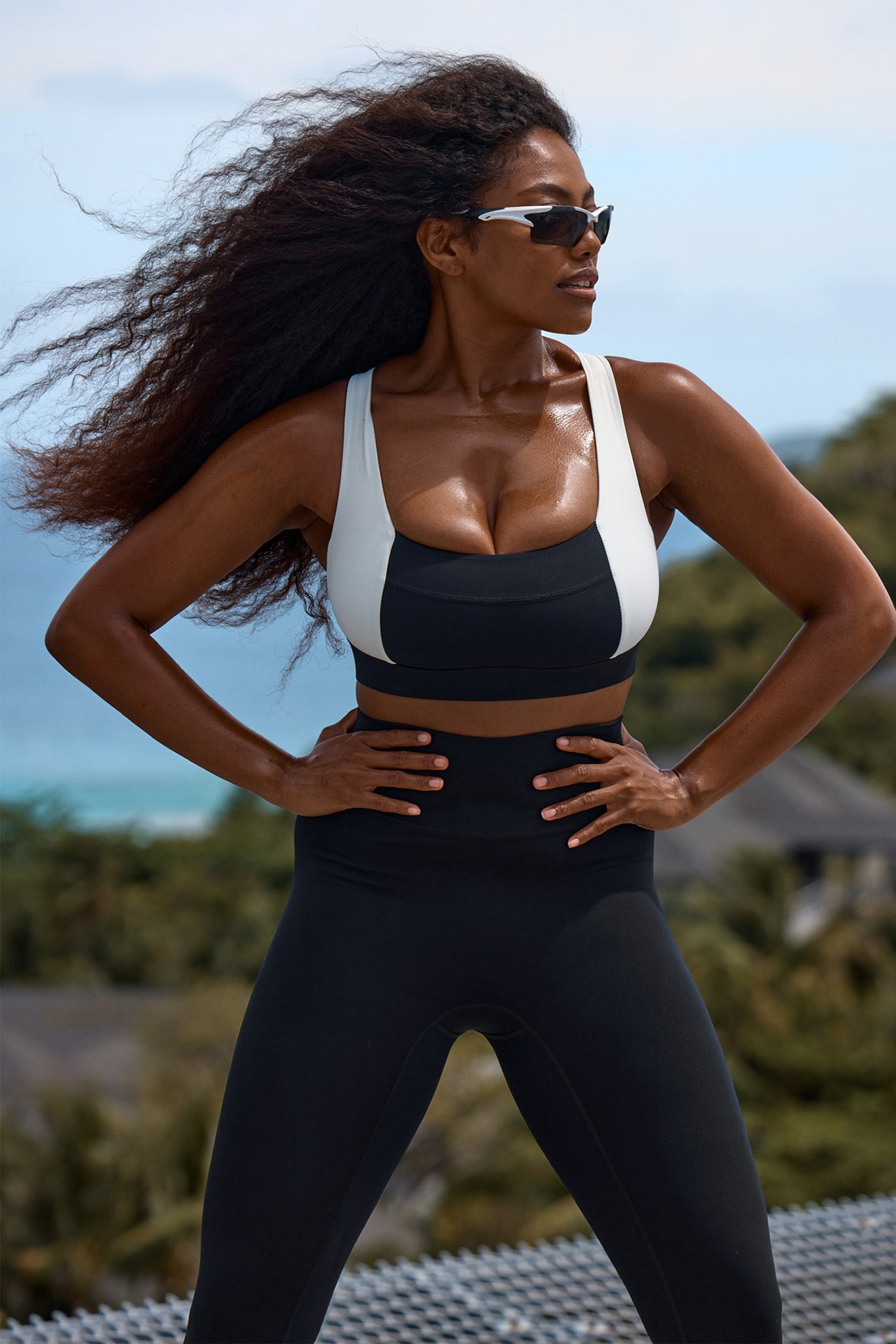 Athletic woman wearing black leggings and a two-tone sports bra, hands on hips, hair blowing with ocean behind her.