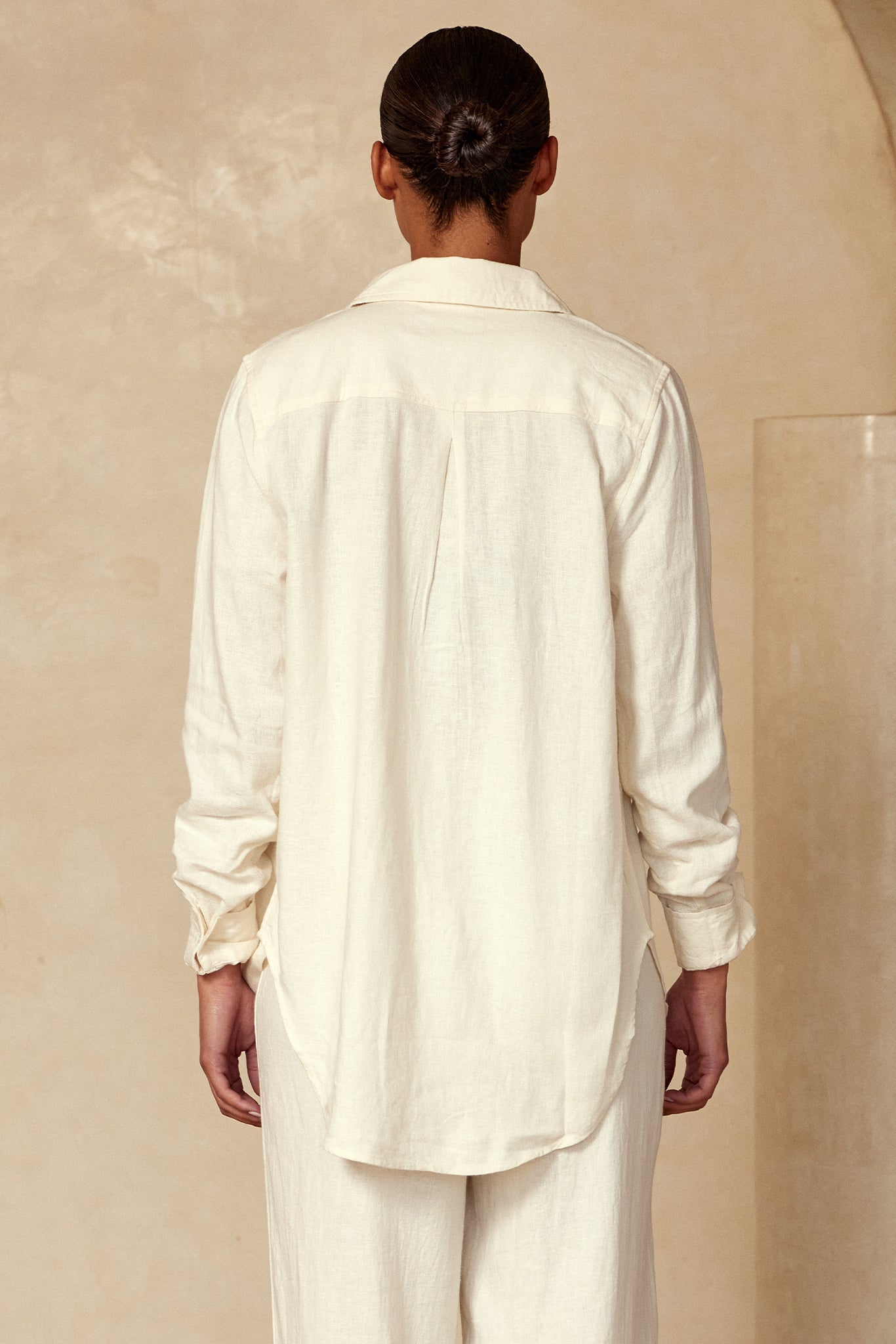 Back view of person in a loose cream linen long‑sleeve shirt and matching wide‑leg pants, hair in a bun, beige backdrop.