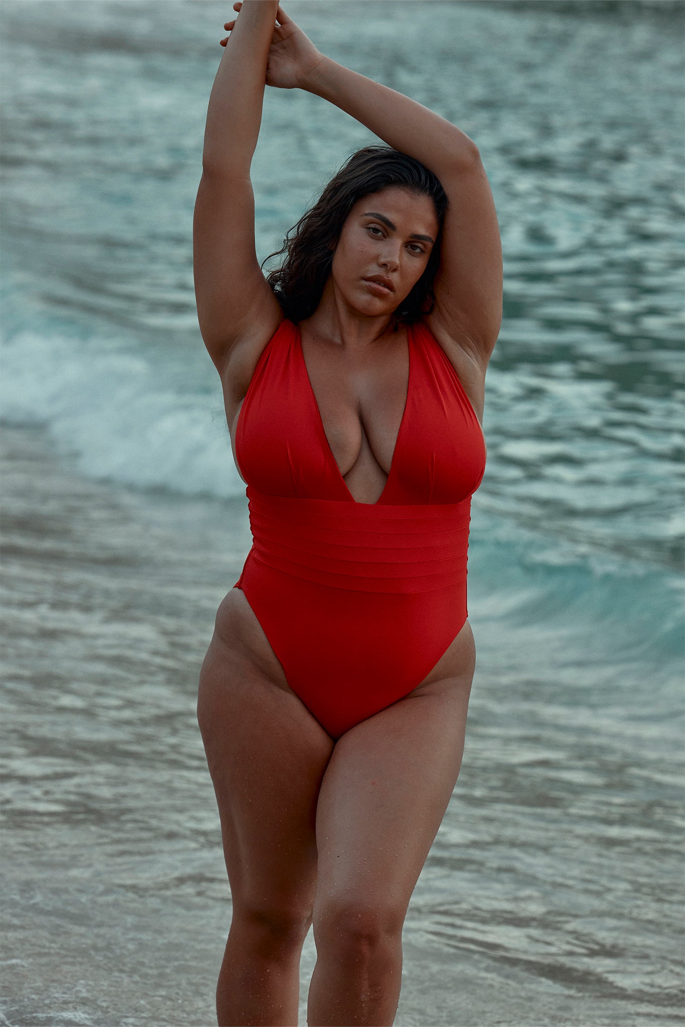 Woman standing waist-deep at the shore in a red one-piece swimsuit with arms raised above her head.