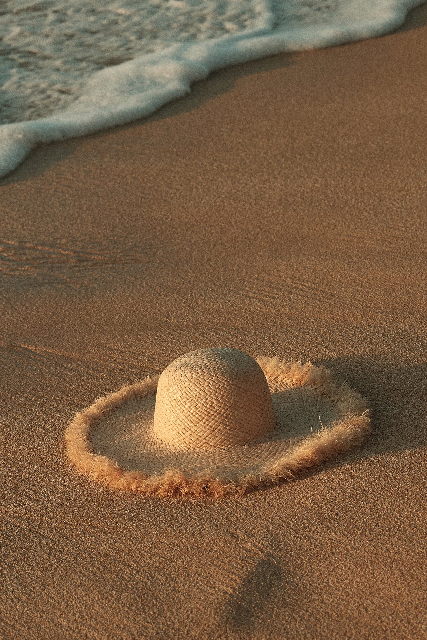 Straw sun hat with frayed brim resting on wet sand near a foamy shoreline
