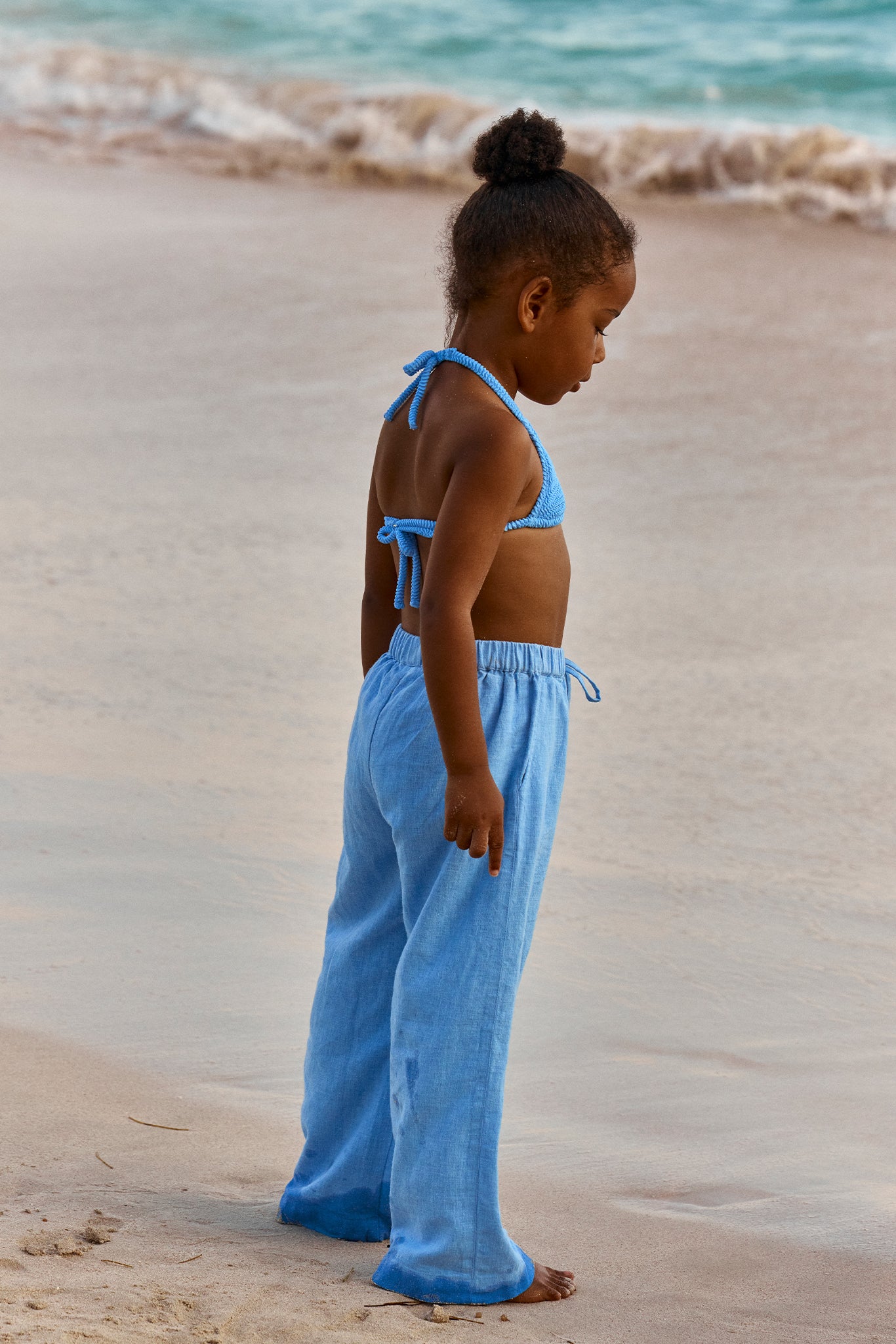 Young girl standing barefoot at water's edge on sandy beach, wearing blue knit bikini top and loose light-blue pants.