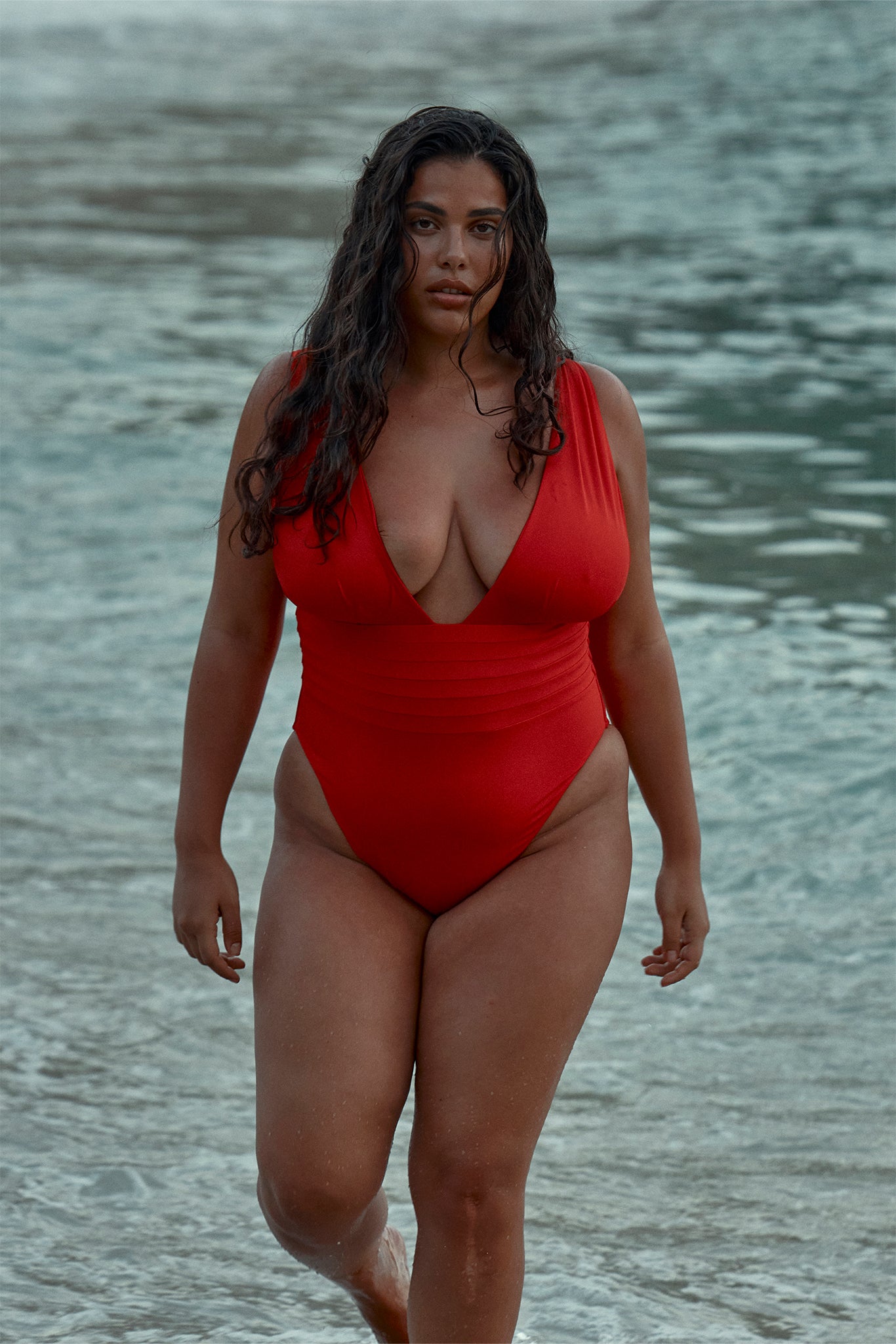 Curvy woman in a red one-piece swimsuit walking out of the ocean with wet hair and neutral expression.