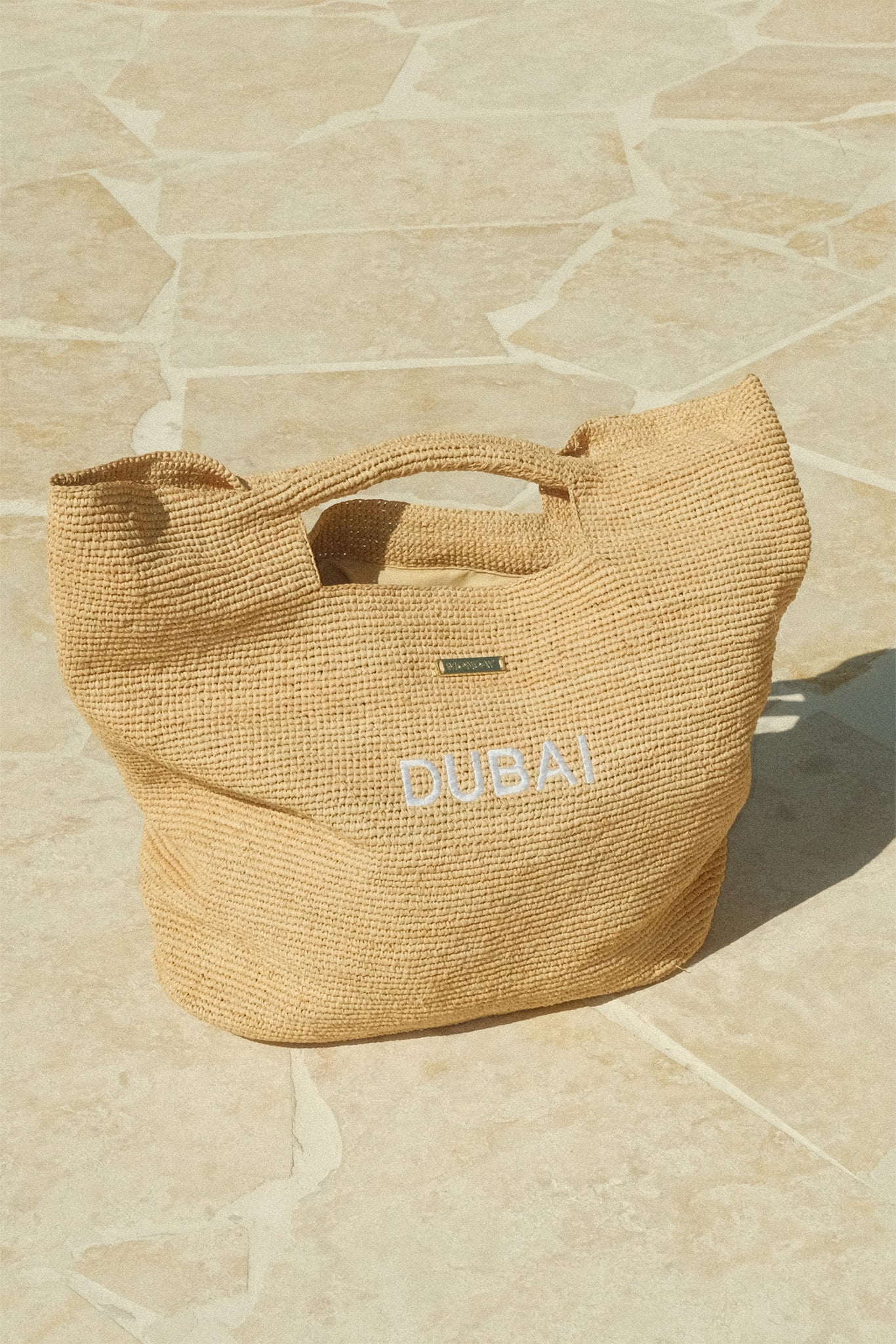 Woven straw tote bag embroidered DUBAI on pale stone-tiled floor