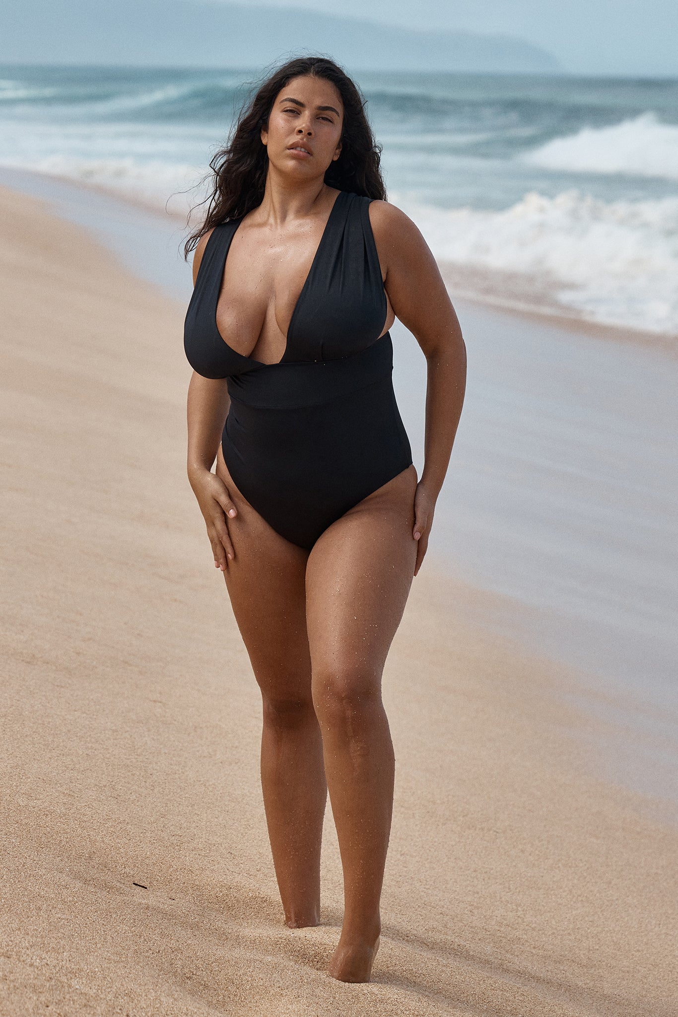 Woman in black one-piece swimsuit walking on a sandy beach with ocean waves in the background.