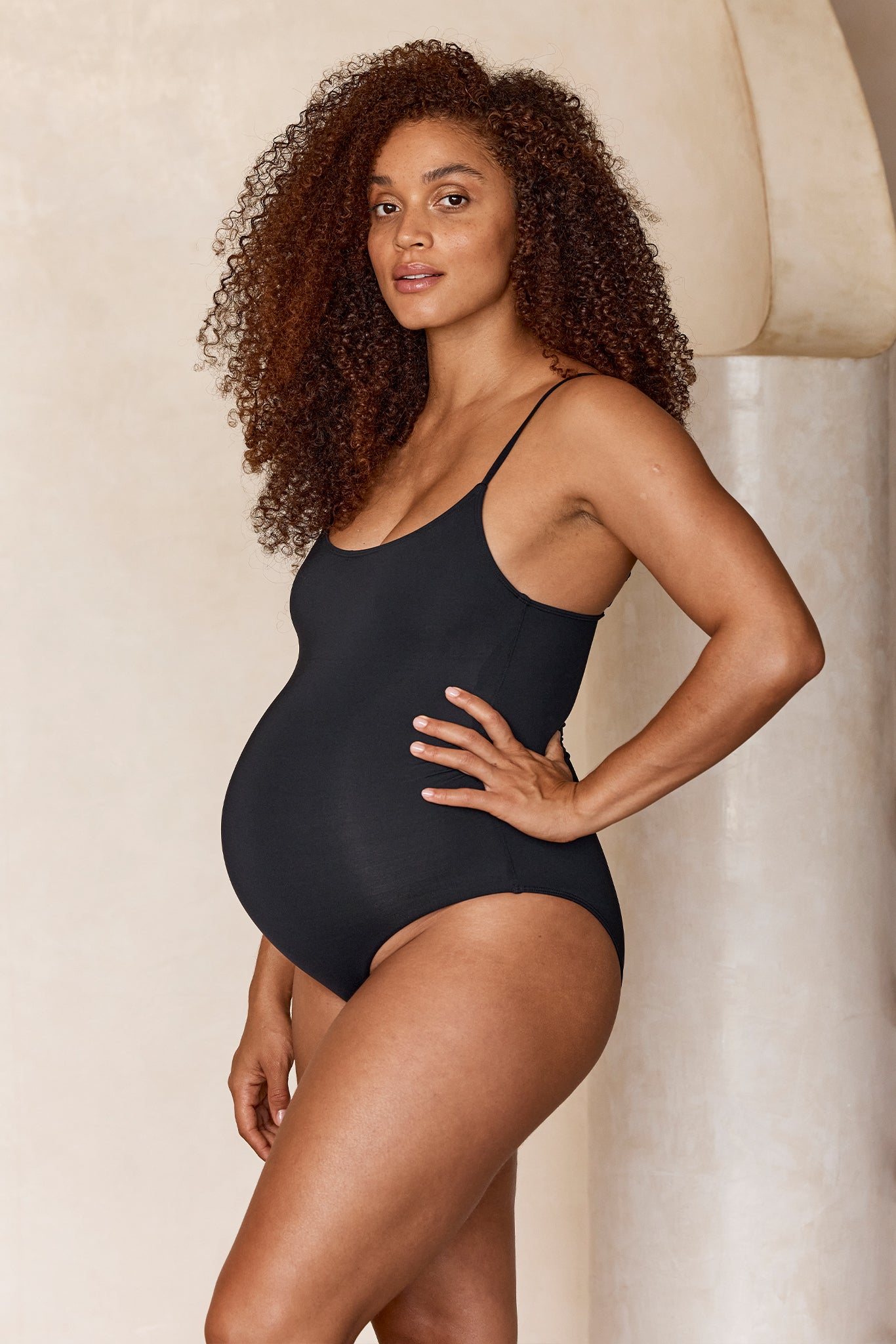 Pregnant woman in a black one-piece bodysuit standing in profile with hand on hip, curly hair, neutral background.