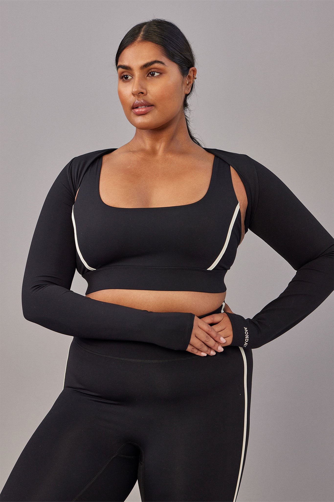 Model in black crop sports bra, long-sleeve shrug, and high-waist leggings with white piping; MONDAY on wrist cuff.