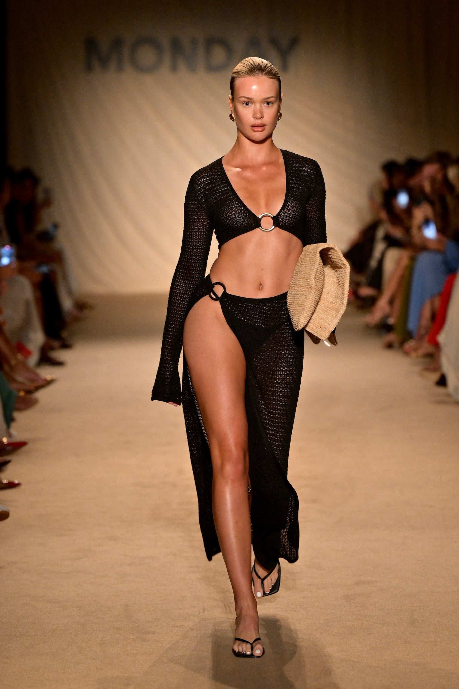 Runway model wearing black knit two-piece and long cover-up holding a straw hat; backdrop reads 'MONDAY'.