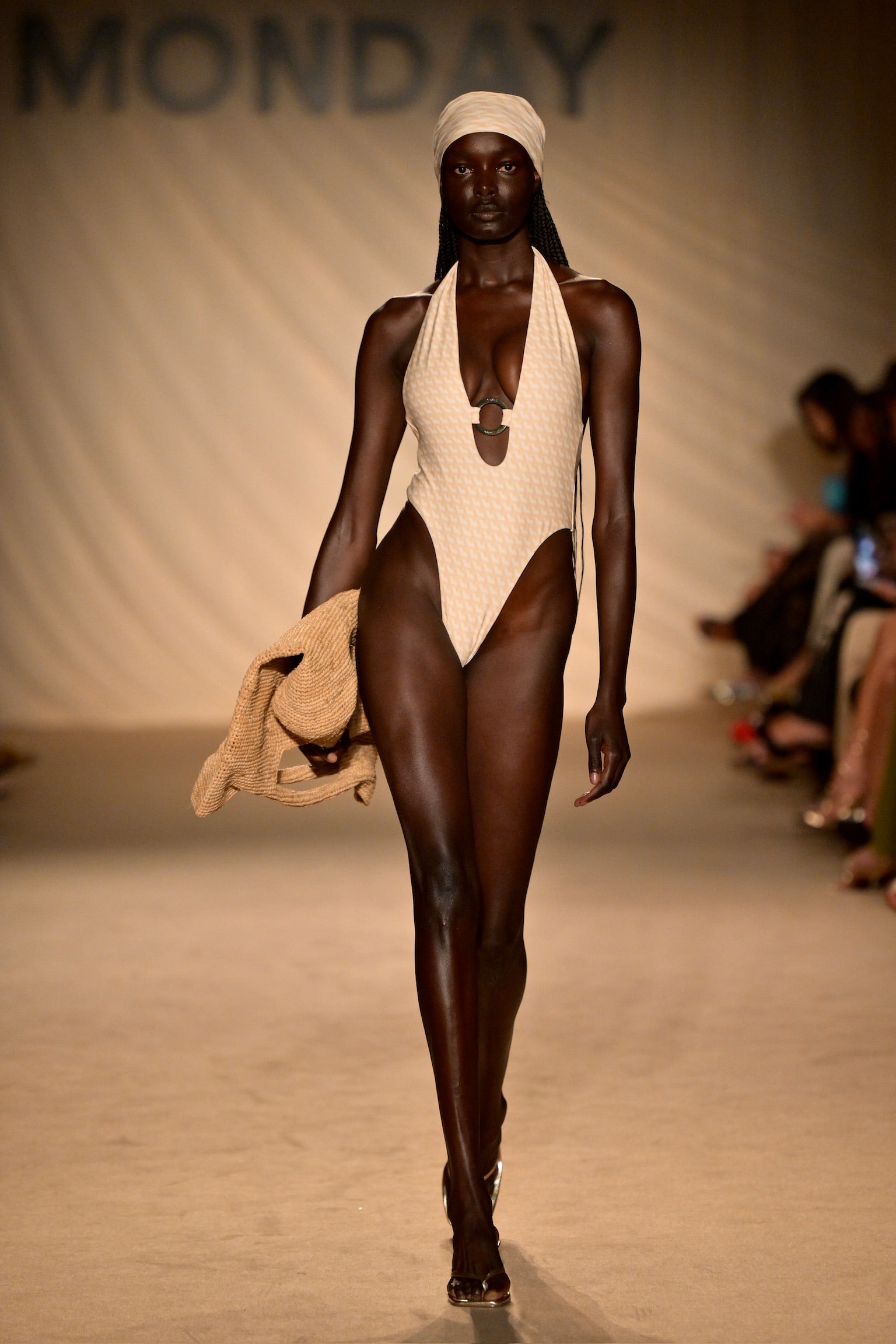Dark-skinned runway model in beige high-cut swimsuit and headscarf holding woven jacket; MONDAY on backdrop.