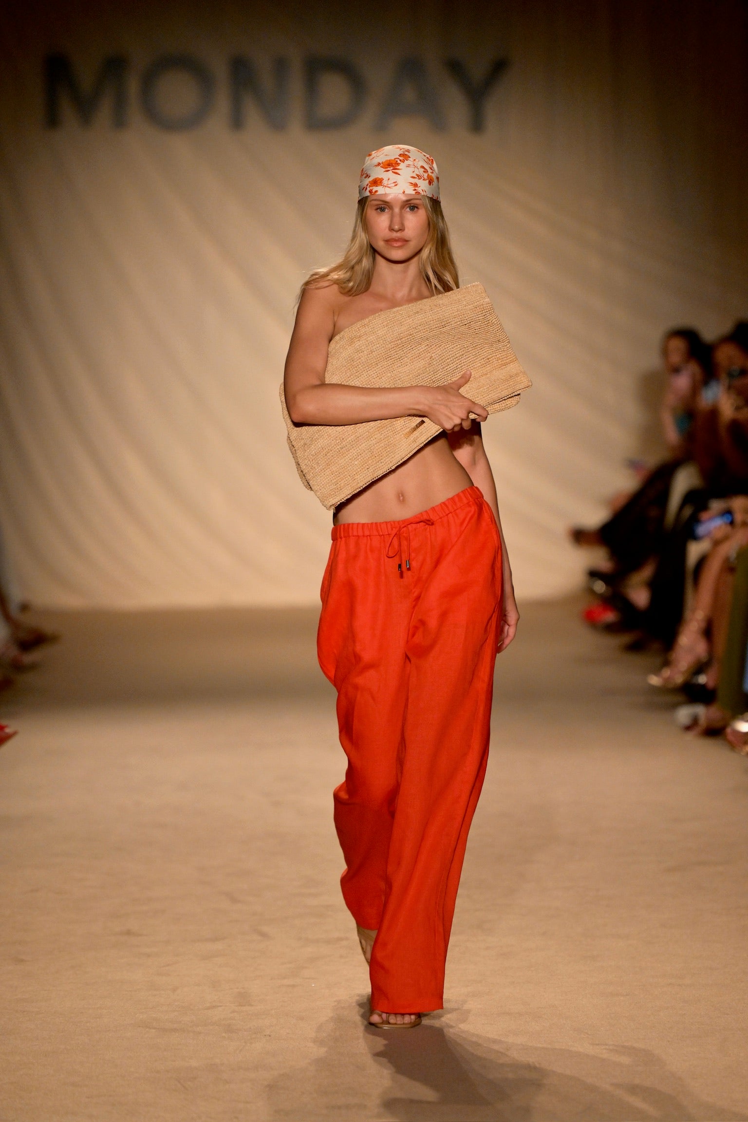 Runway model in orange wide-leg pants and floral headscarf holding a woven clutch; backdrop reads MONDAY.