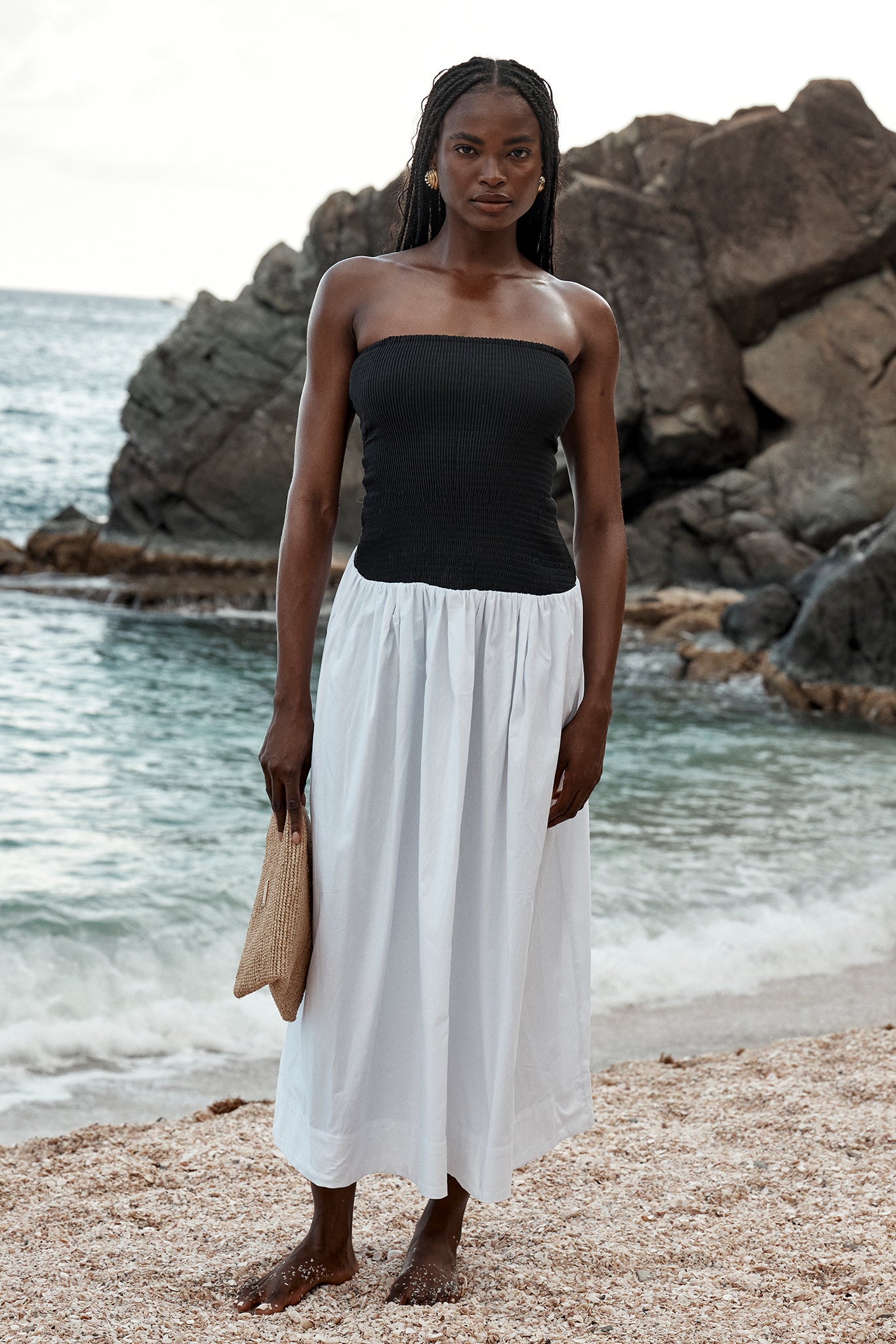 Woman standing barefoot on a rocky beach wearing a strapless black top and white midi skirt, holding a woven clutch