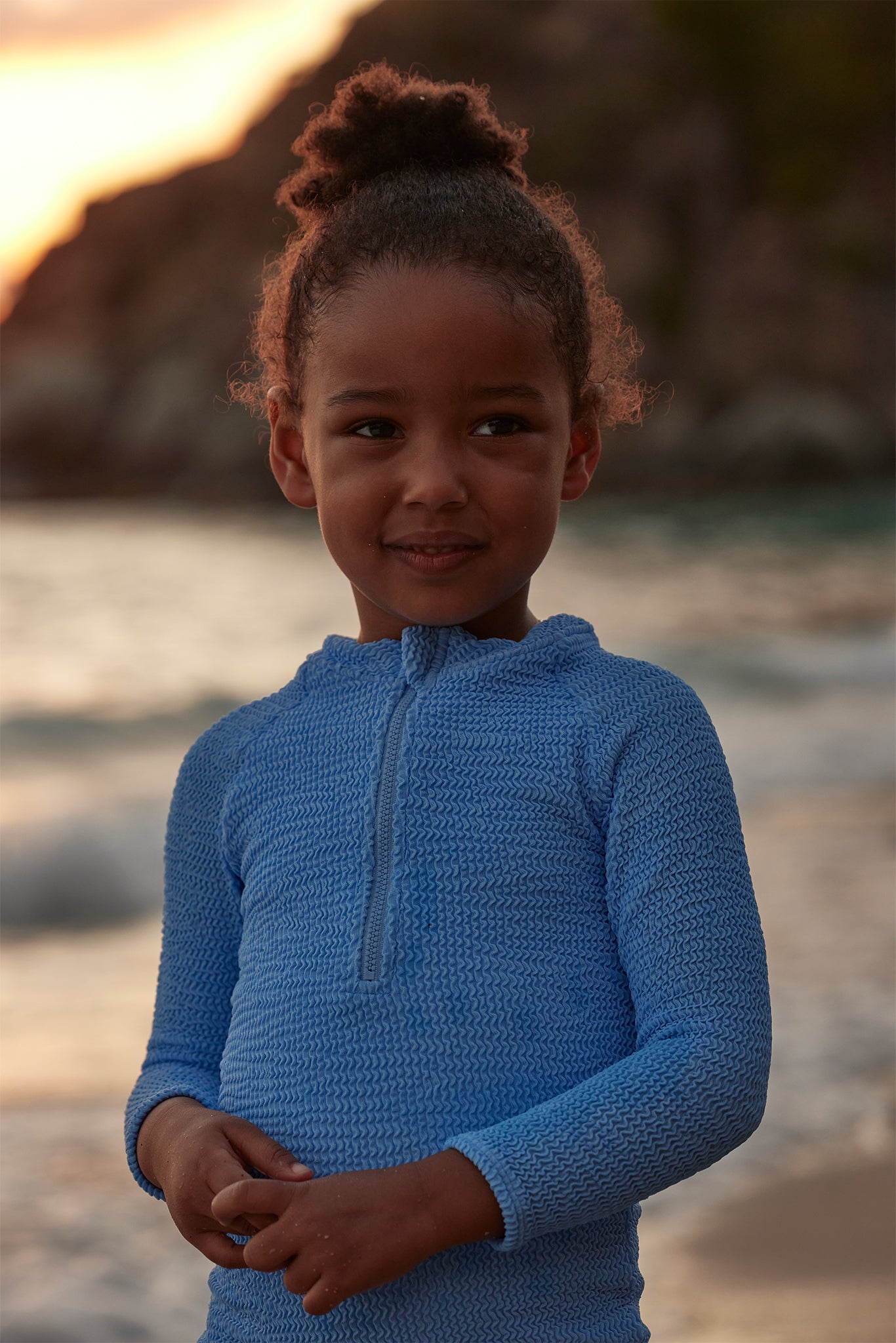 Young girl standing on a beach at sunset wearing a textured light-blue zip-neck top, hands clasped