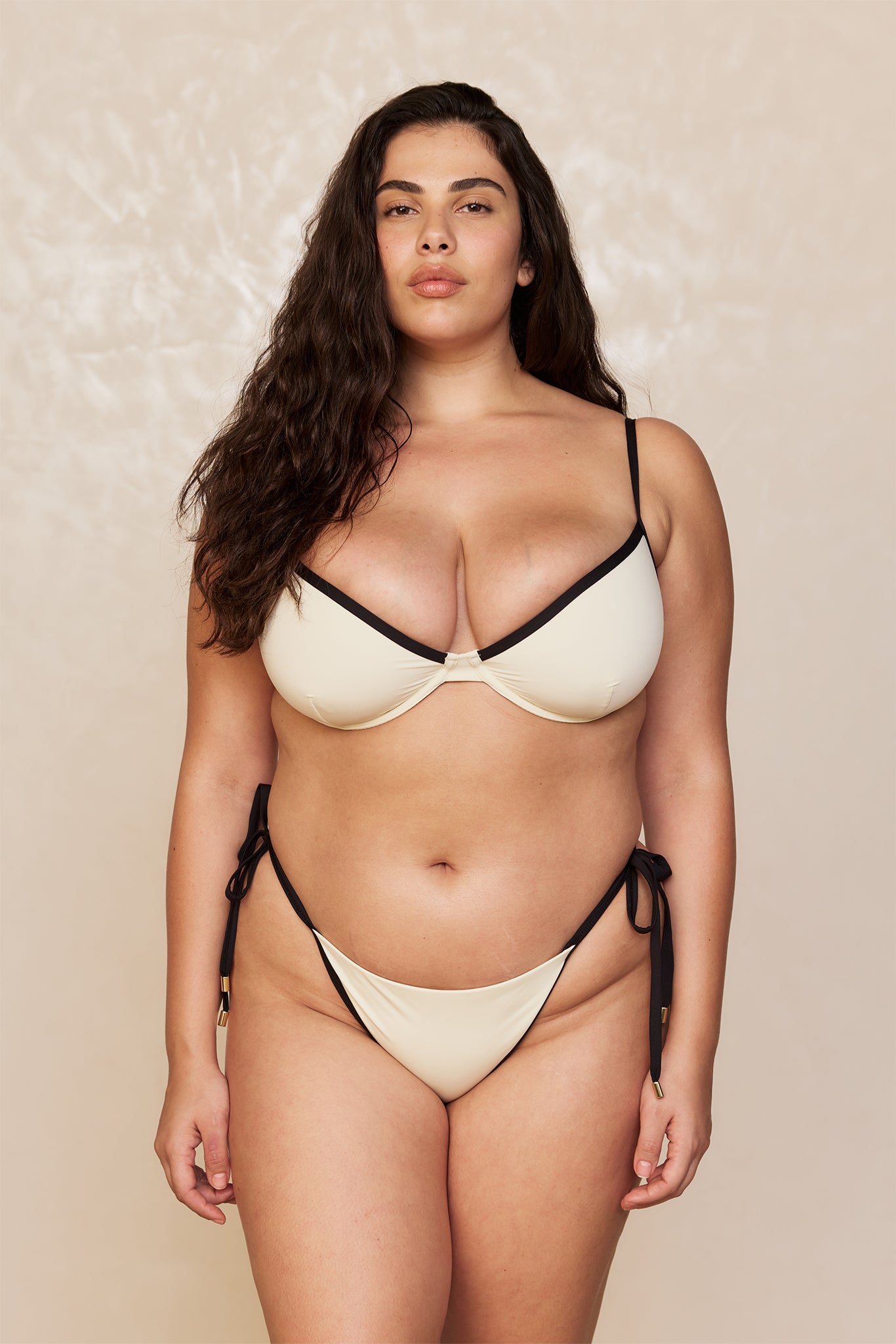 Curvy model facing camera wearing cream bikini with black trim and tie-side bottoms, long wavy dark hair, neutral backdrop.