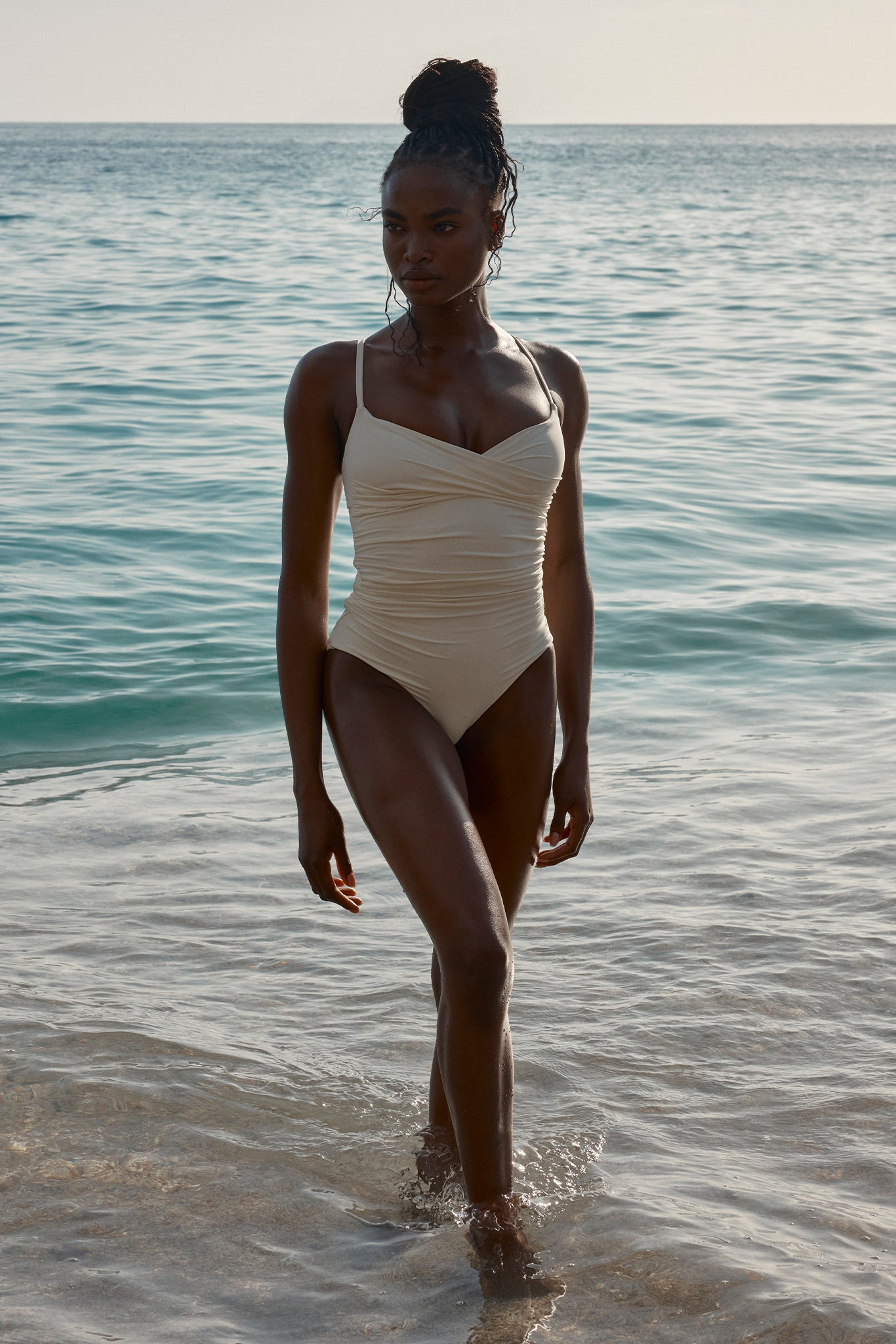 Person in a beige one-piece swimsuit walking out of shallow sea with hair in a bun and sunlit waves behind.