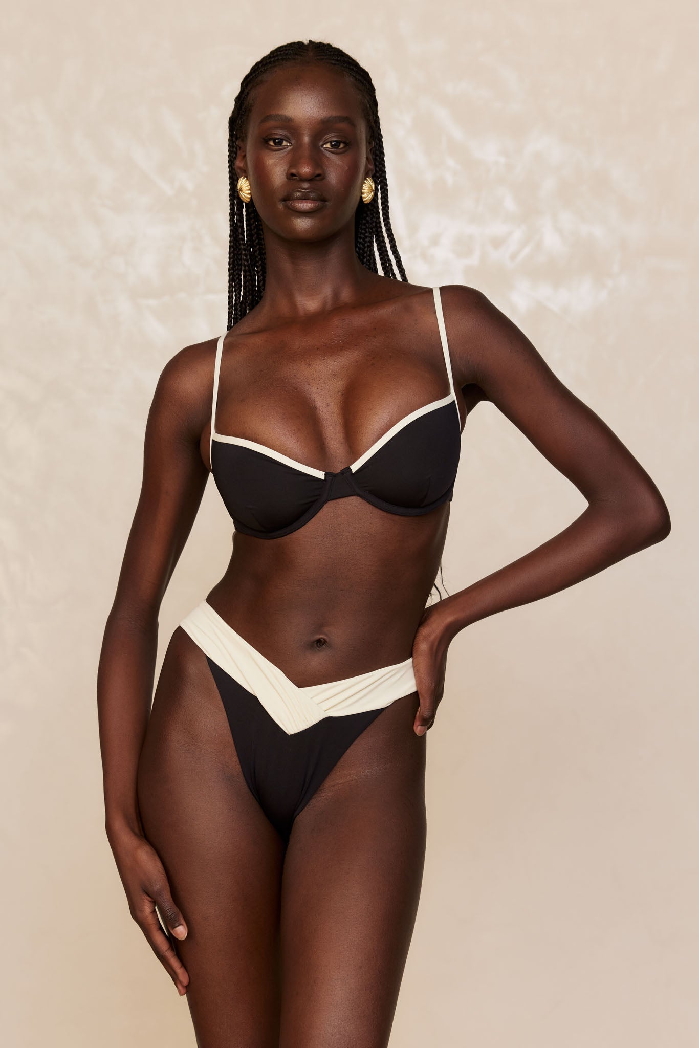 Dark-skinned model with braids and gold earrings wearing black bikini with cream trim, hand on hip, beige backdrop.