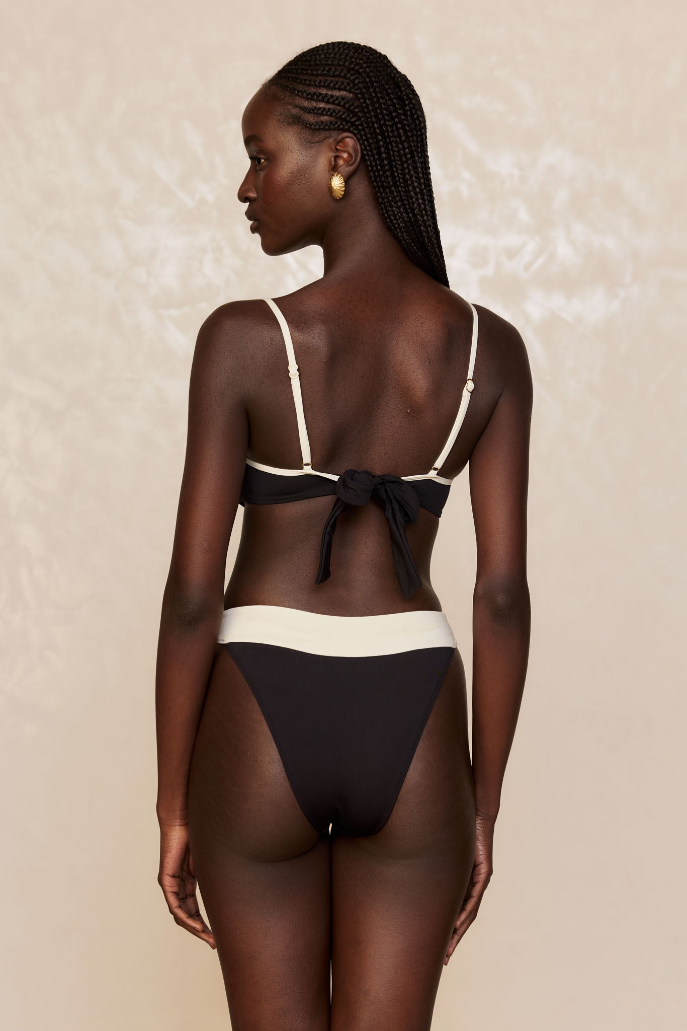 Rear view of a model wearing a black-and-cream bikini with tied-back top, high-cut bottoms, braided hair and gold shell-shaped earrings.