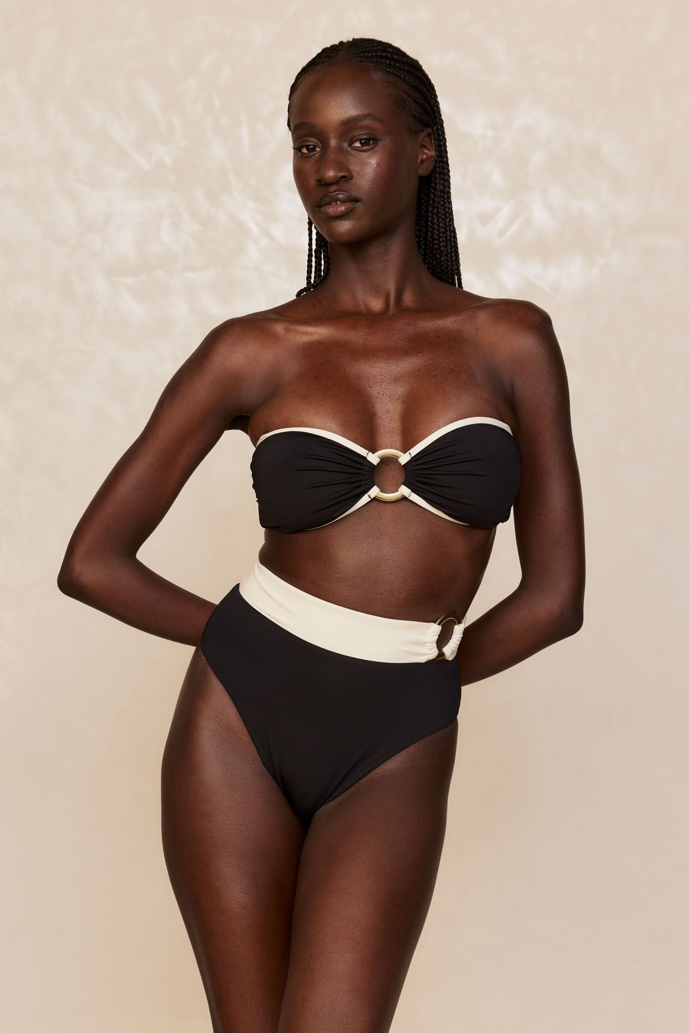 Woman wearing black bandeau bikini with cream trim and ring accents, high-waist bottoms, hands behind back, neutral backdrop.