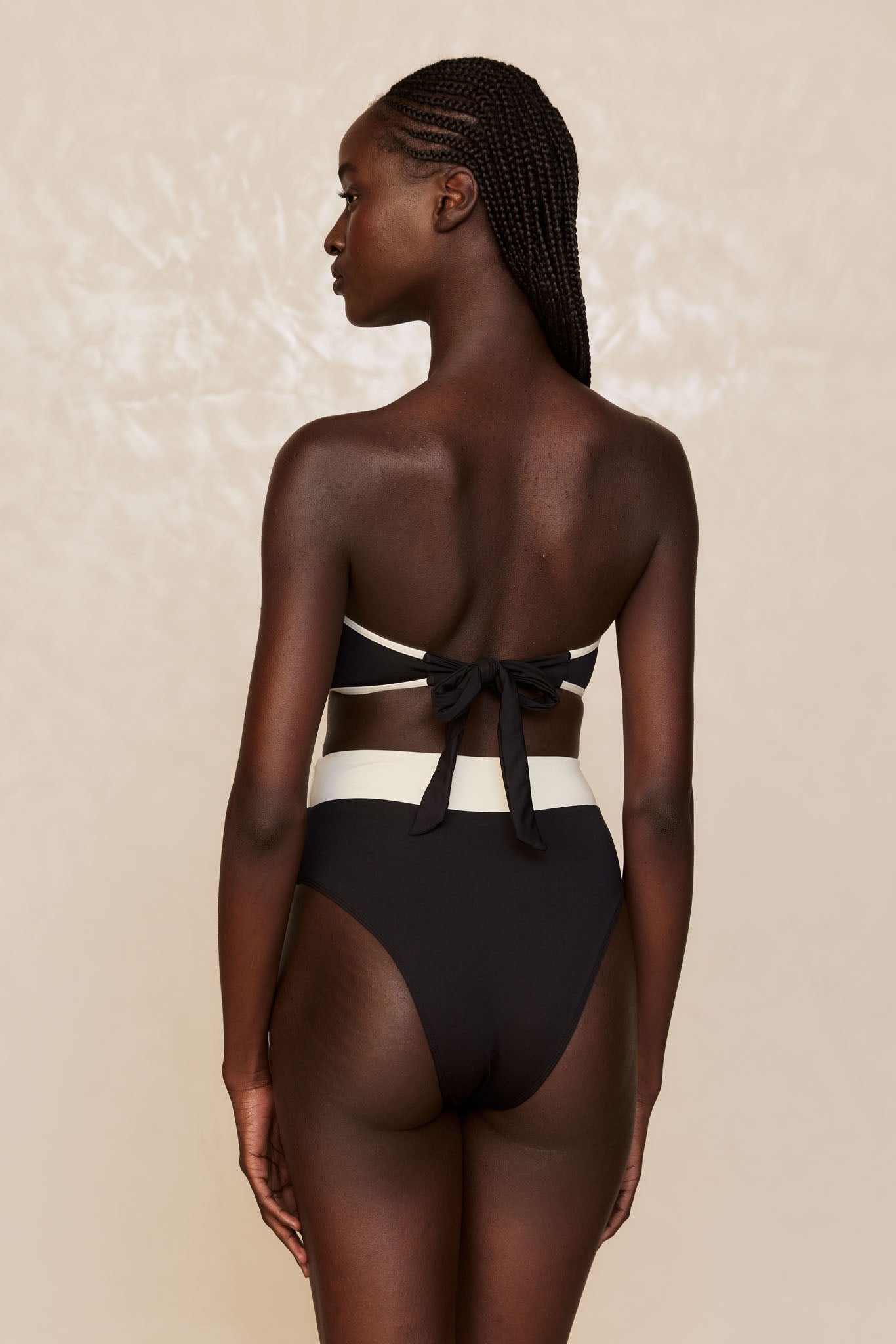 Back view of model with braided hair wearing black high-waist bikini bottoms and a bandeau top tied in a bow