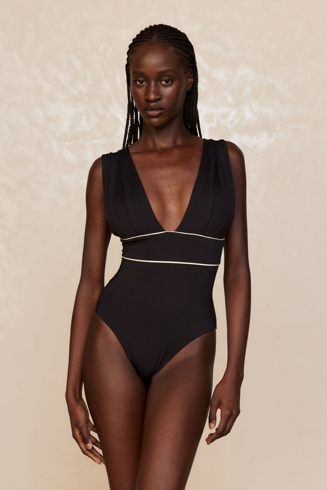 Woman with braided hair wearing a black one-piece swimsuit with deep V-neck and white piping, on beige background.