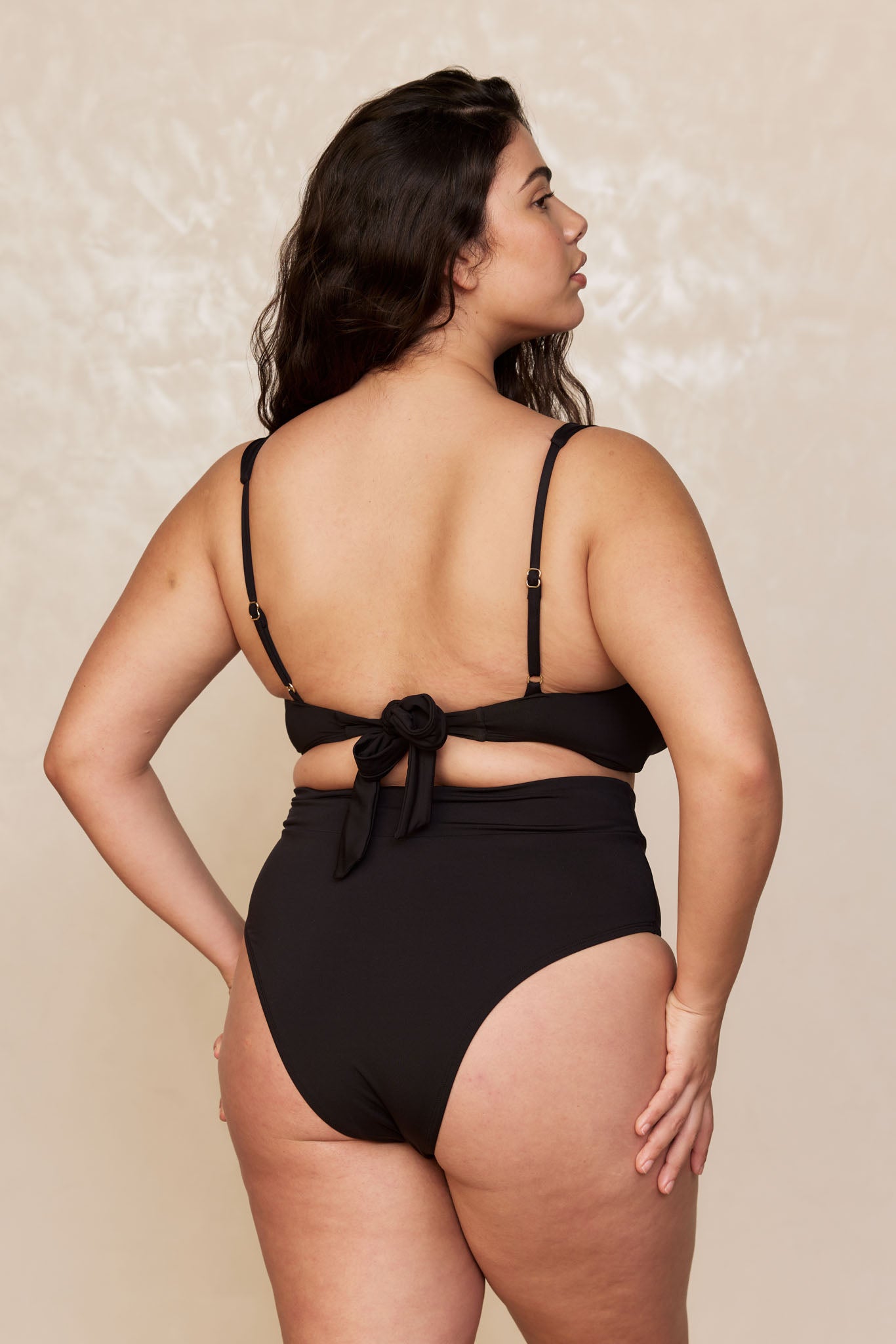 Curvy woman shown from back wearing black high-waist bikini bottoms and a tied-back bikini top, head turned to the right.