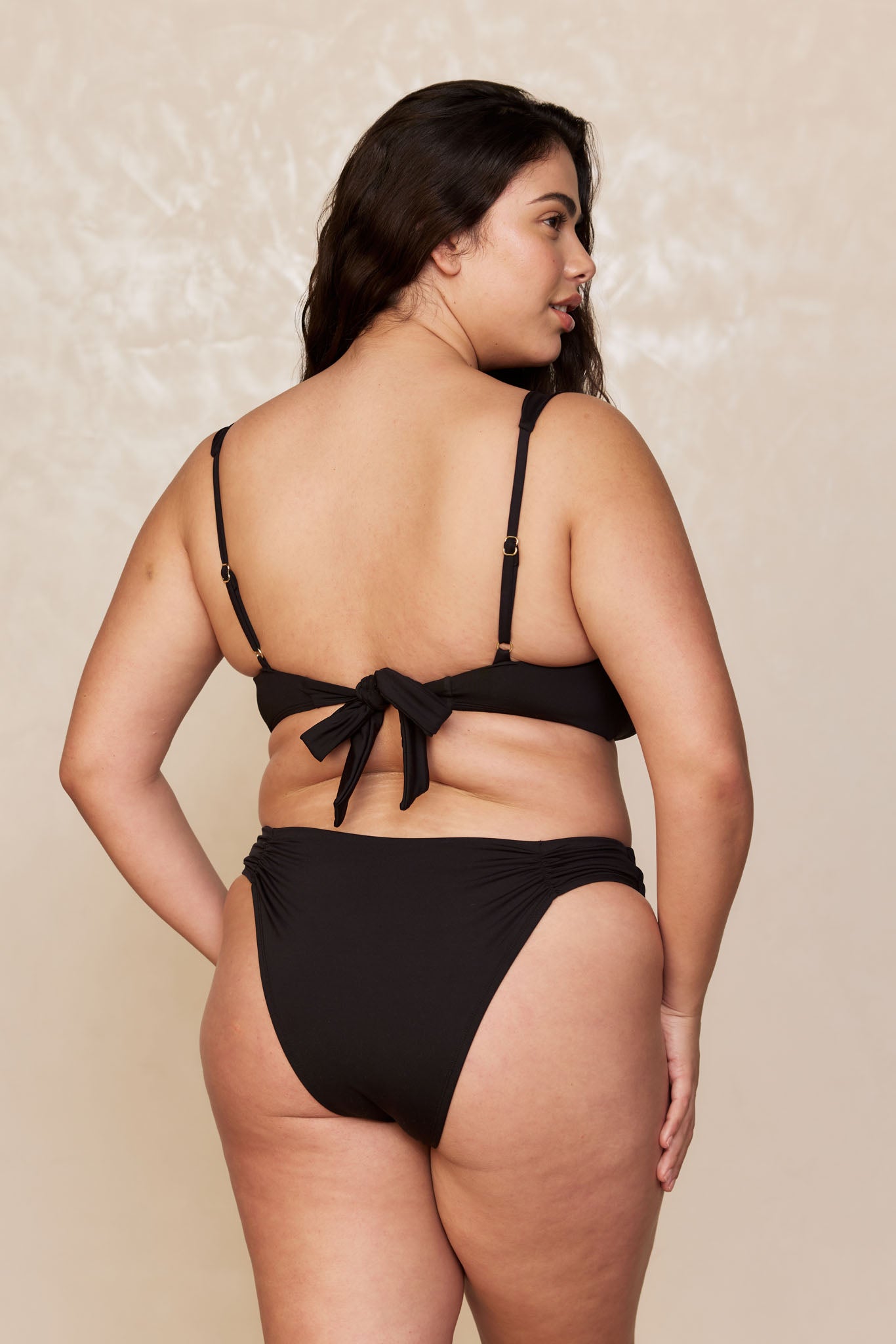 Rear view of a plus-size woman wearing a black bikini with a tied back and high-cut bottoms on a neutral backdrop.