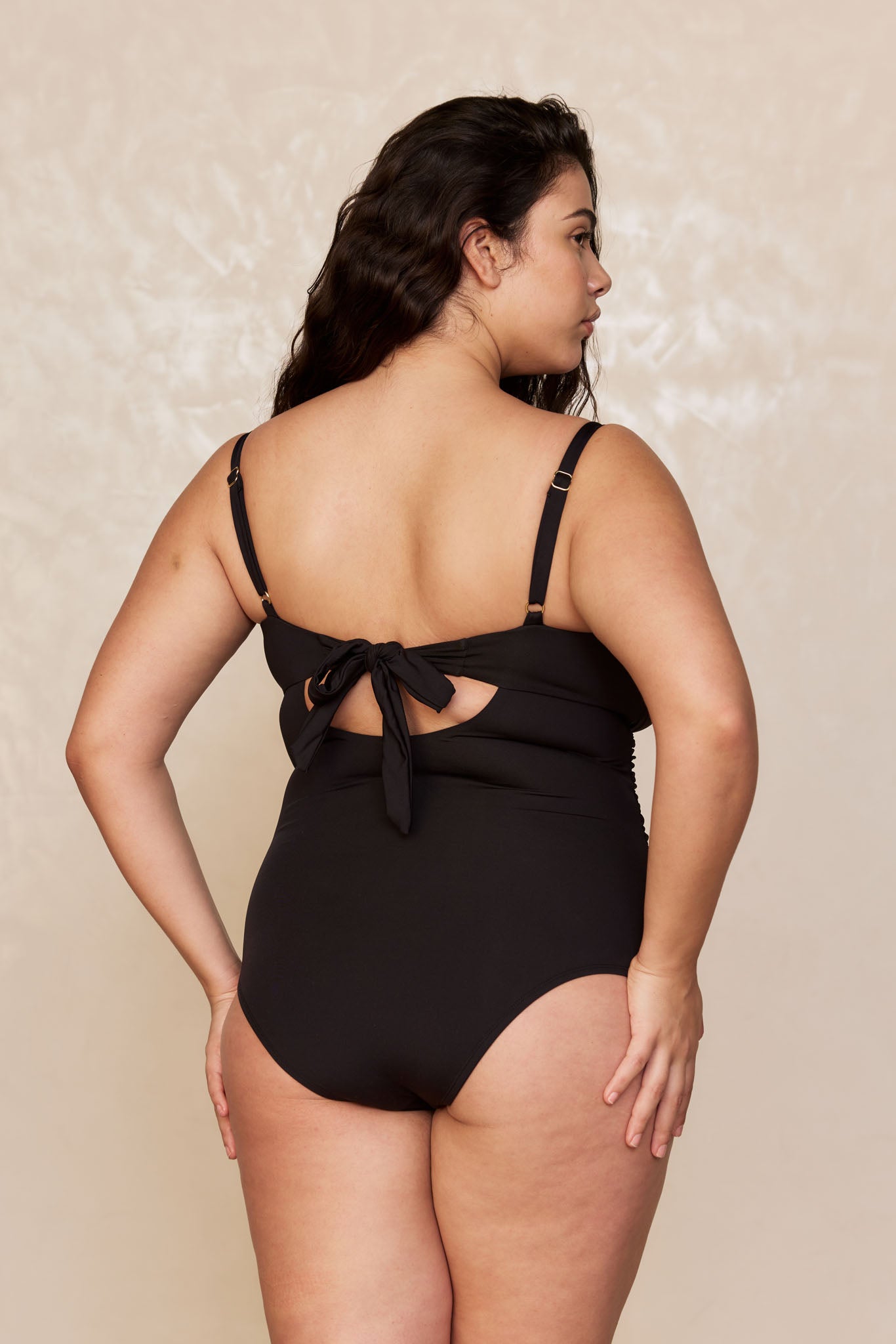 Back view of woman wearing black one-piece swimsuit with adjustable straps and tied bow at back.