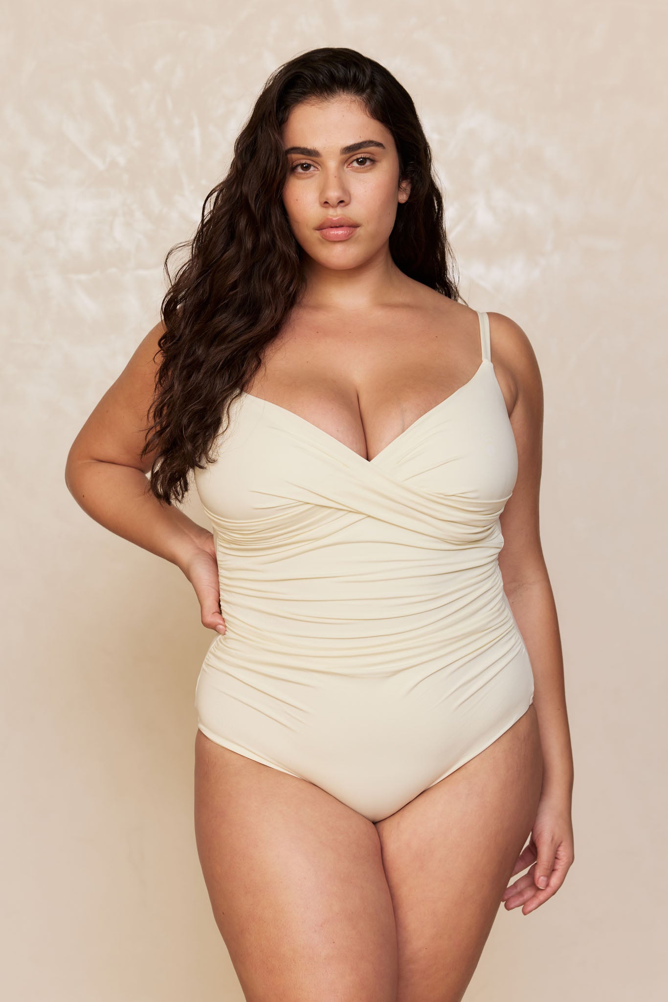 Plus-size model with long dark wavy hair wearing a cream ruched one-piece swimsuit, posing with hand on hip against a neutral background