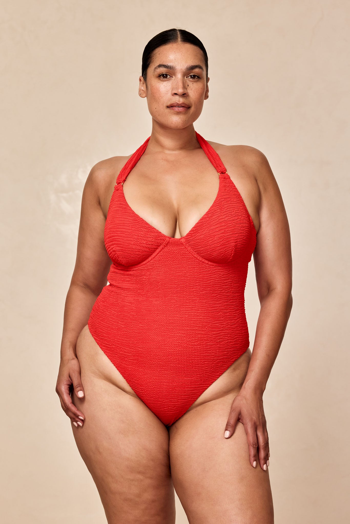 Plus-size woman front-facing in a red textured halter one-piece swimsuit against a beige background.