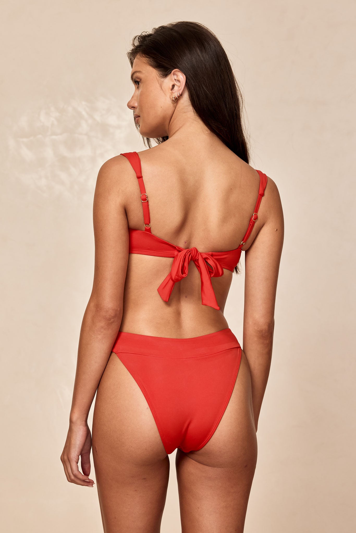 Woman shown from back wearing red bikini with tied bow at back and high-waist bottoms, neutral studio background