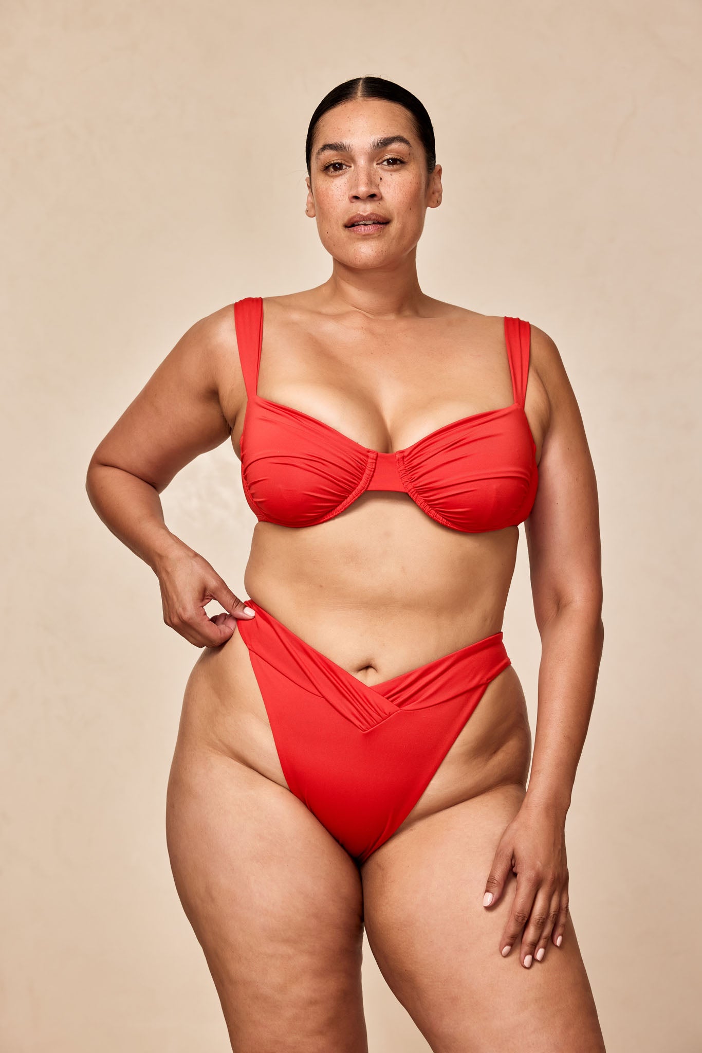 Plus-size woman in red underwire bikini, front view, hand adjusting high-waist bottoms against neutral studio backdrop.
