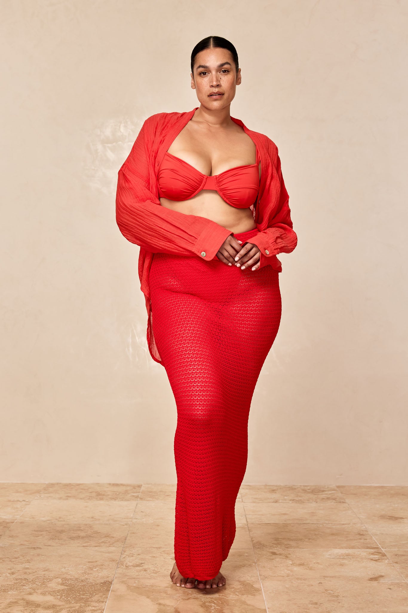 Woman wearing red bikini top, open red shirt and long crochet skirt, standing barefoot on tiled floor against a neutral background.