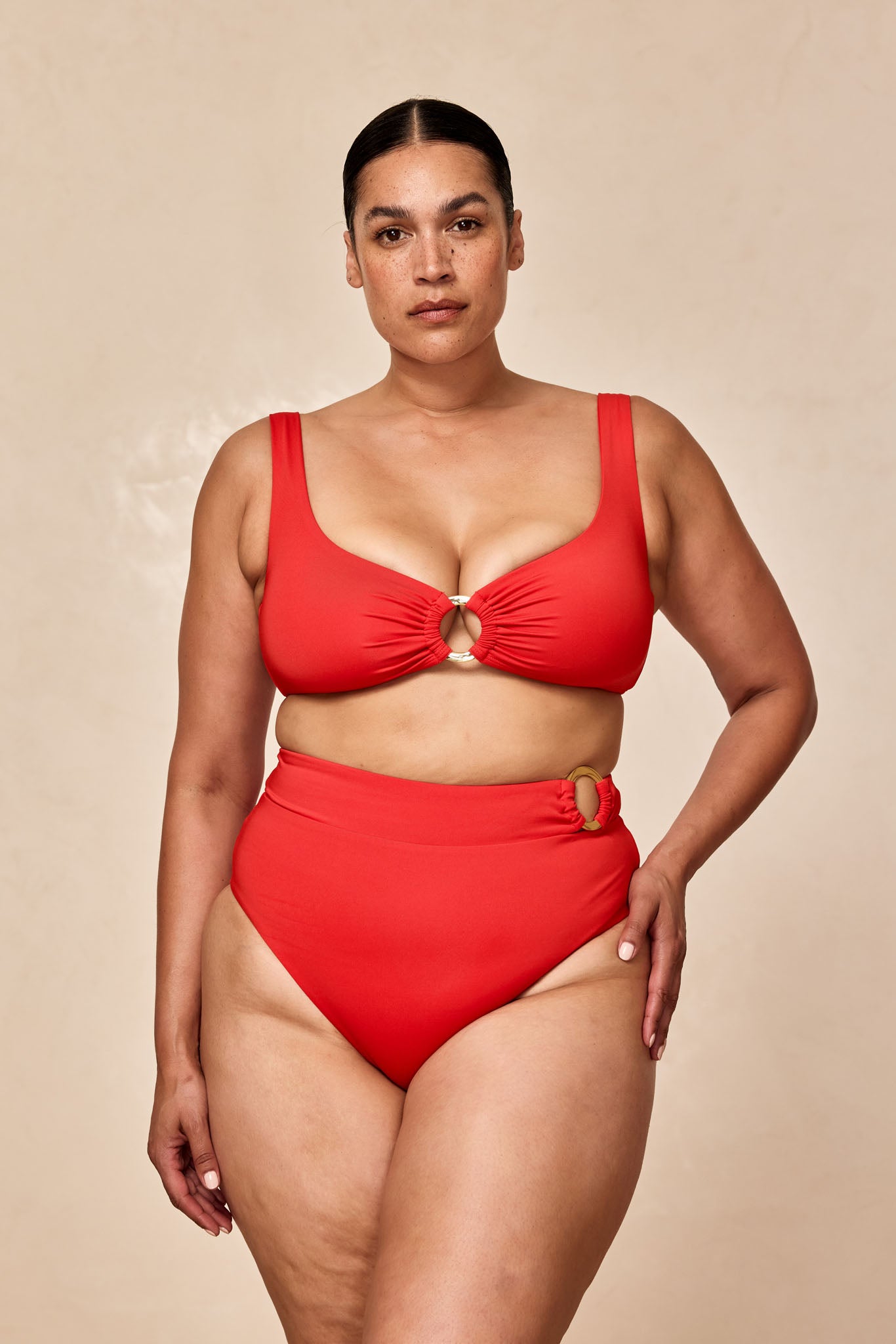 Plus-size woman wearing red two-piece swimsuit with high-waist bottoms and ring detail at center and hip, neutral background.
